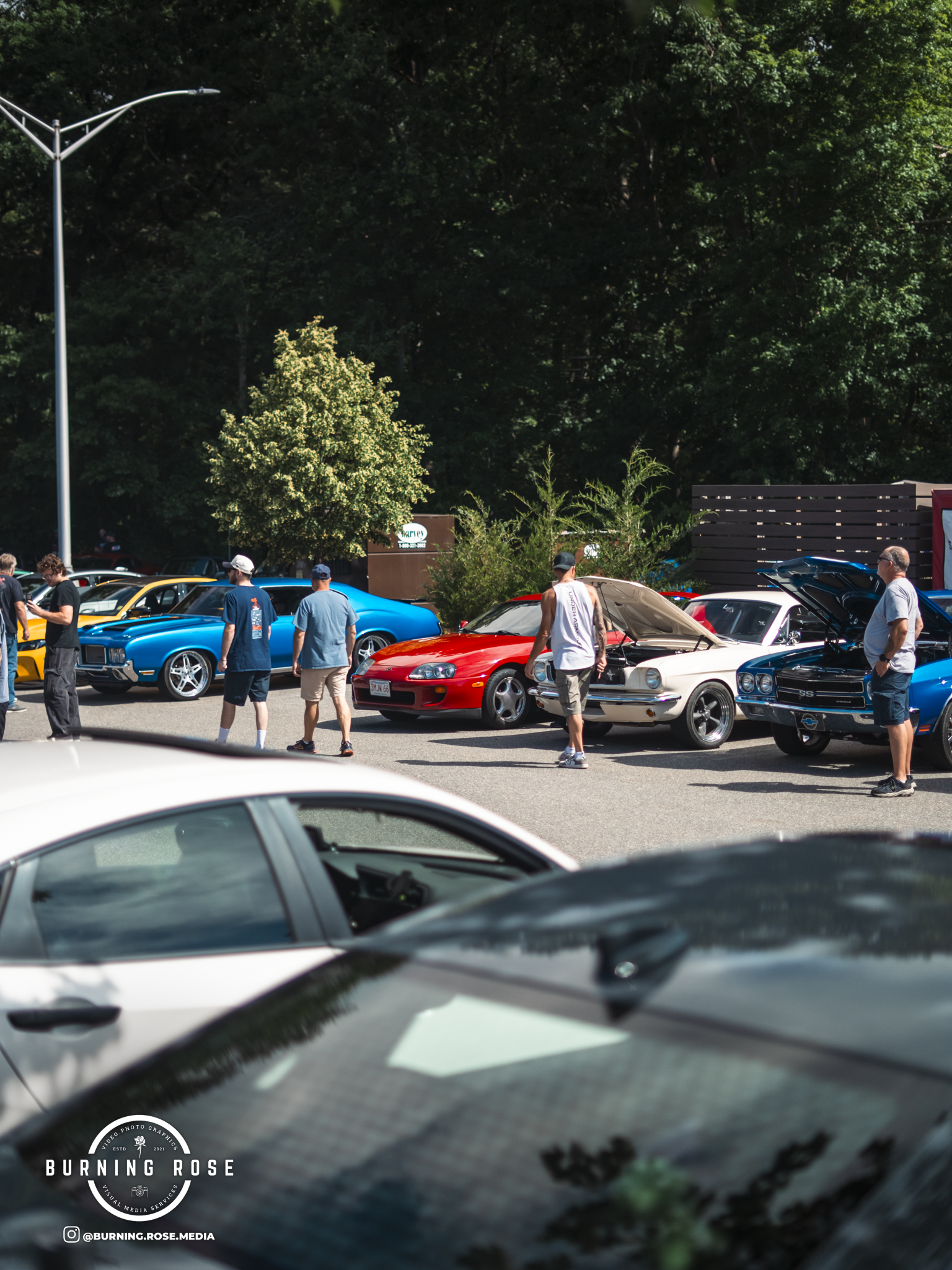 People attending a car show with classic and muscle cars parked outdoors. The scene features various vintage cars, some with their hoods open, and attendees walking around and admiring the vehicles. The background shows trees and a partially cloudy s