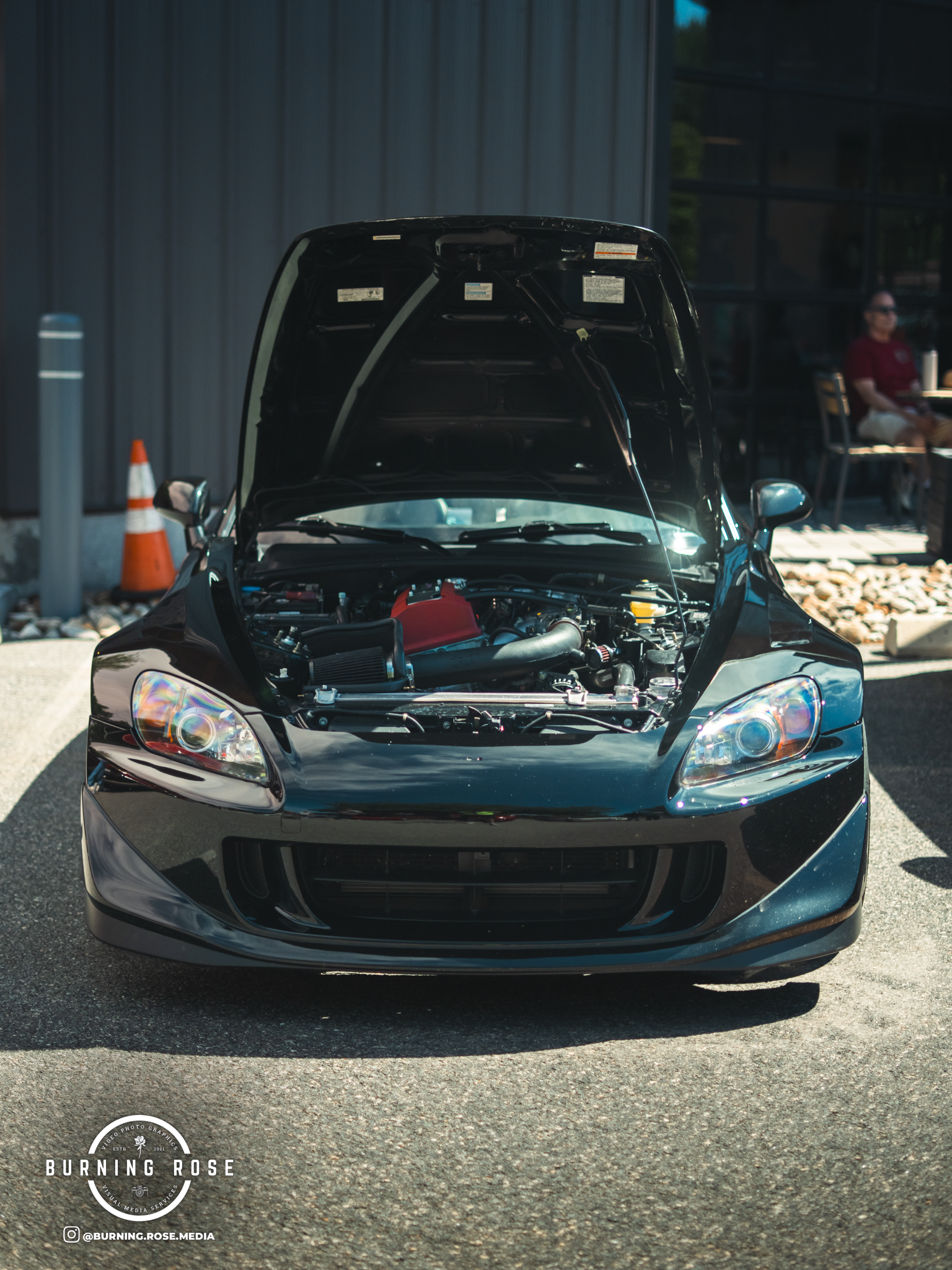 A black sports car with its hood open, showing the engine compartment, parked outdoors on a sunny day. There is a man sitting at a table in the background on the right, wearing sunglasses and a red shirt.