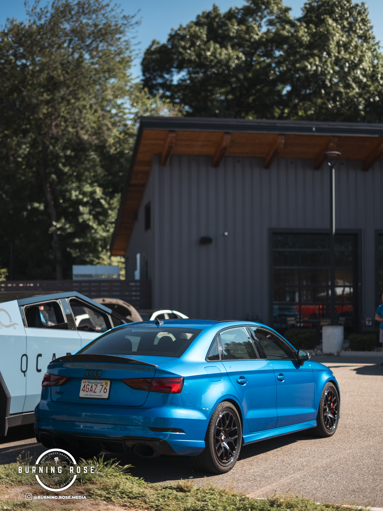 Close-up of a blue Audi car parked outdoors, with other vehicles nearby, in front of a building with a metal exterior and trees in the background.