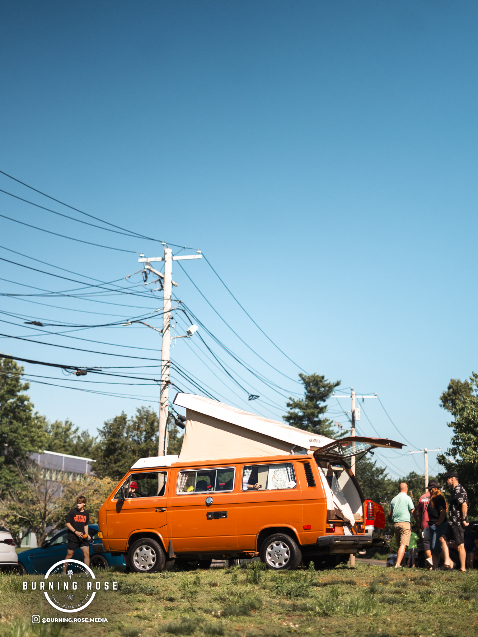 An orange van with a pop-up roof is parked on a grassy area. Several people are walking around and socializing nearby. A utility pole with wires and a clear blue sky are visible in the background.