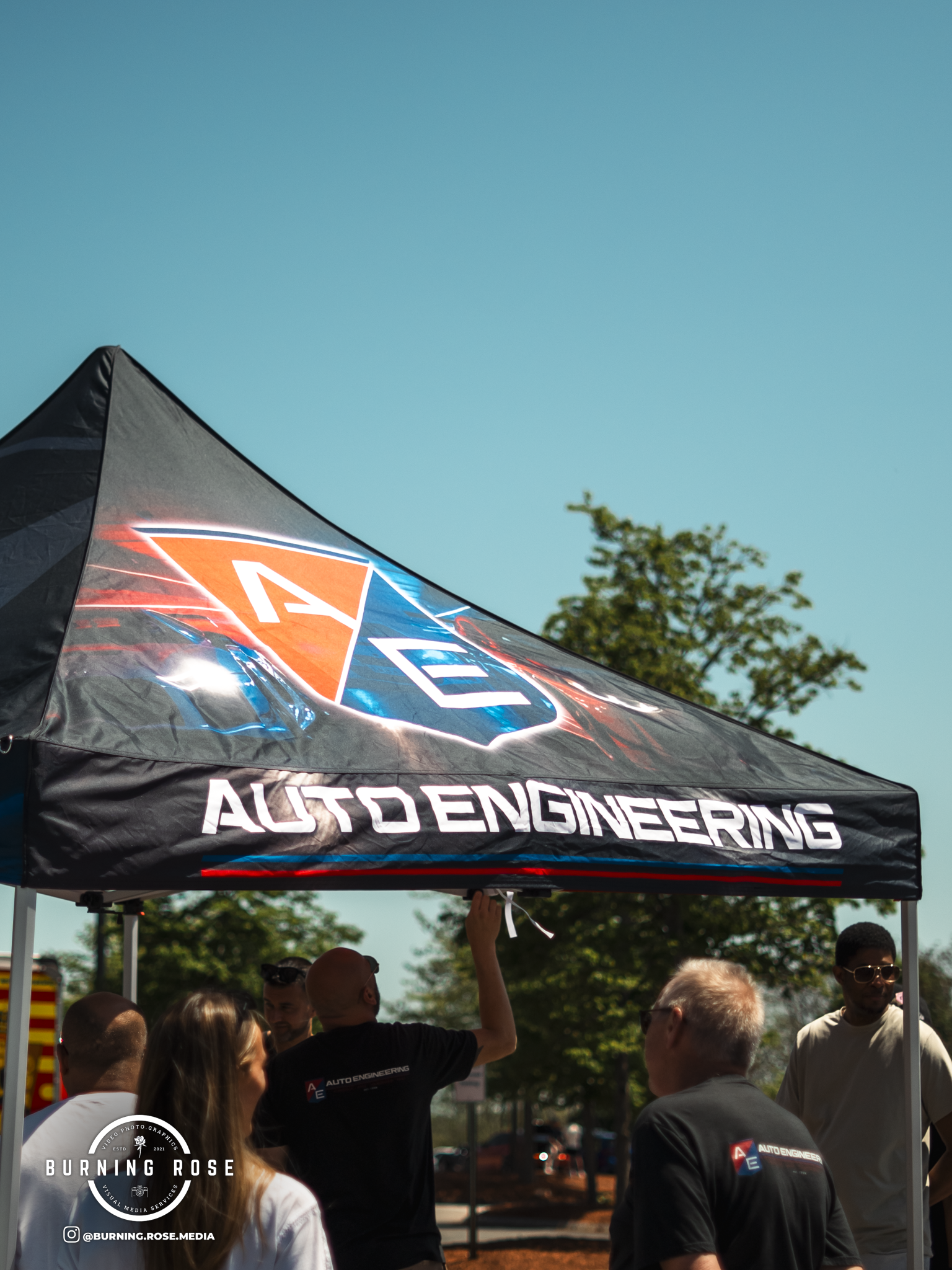 A group of people gathered at an outdoor event under a black canopy with the words "Auto Engineering" printed on it. The sky is clear and blue, and some trees are visible in the background.