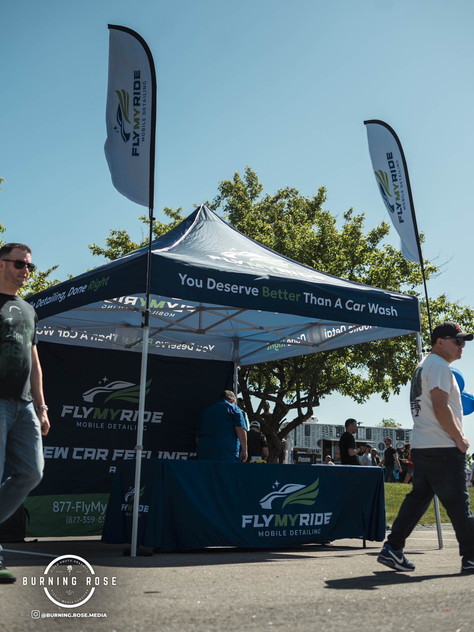 A promotional booth for FlyMyRide Mobile Detailing with two tall flags and a tent set up outdoors on a sunny day, with people walking nearby and trees in the background.