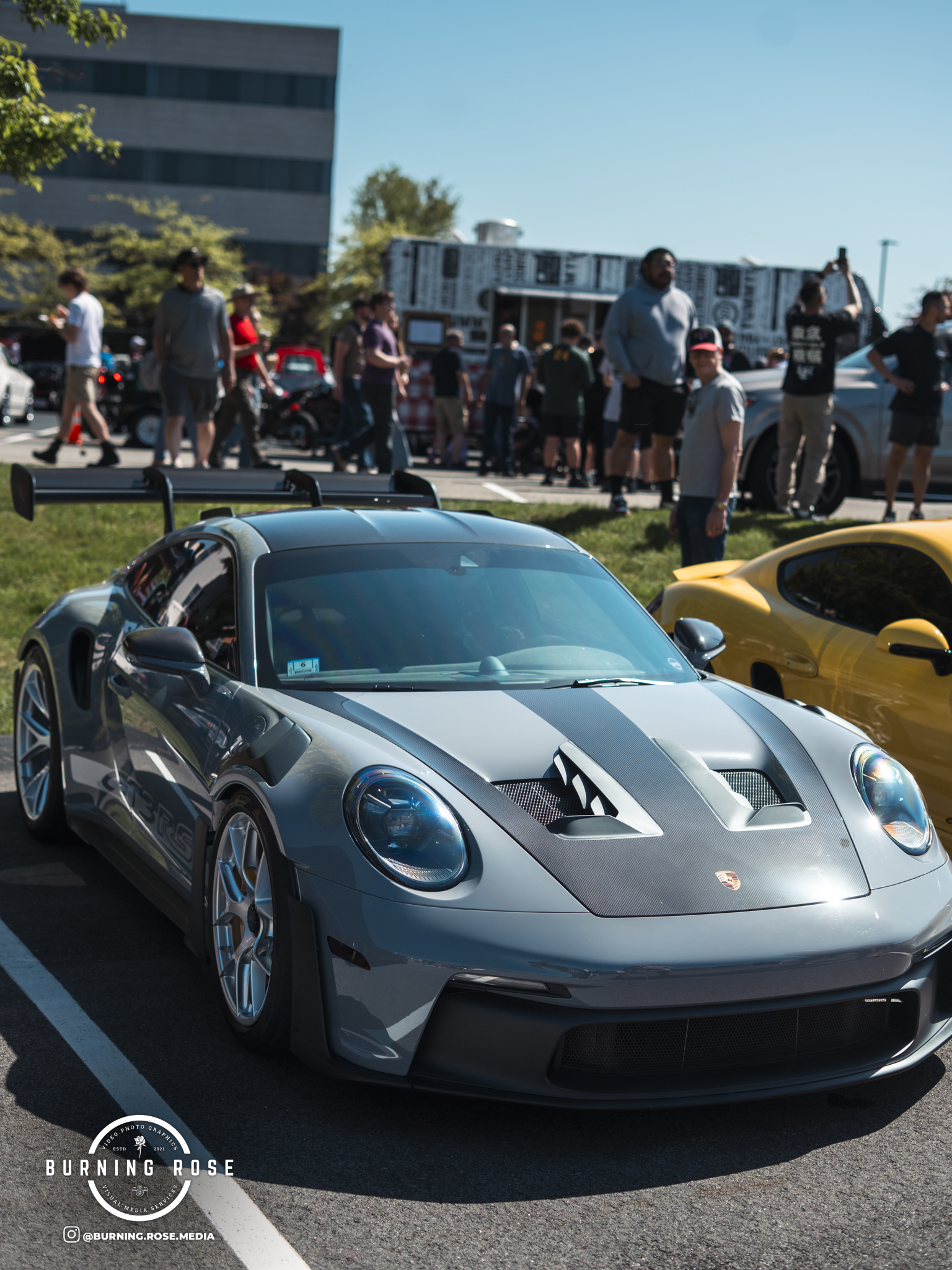 A silver sports car with a carbon fiber hood parked in a parking lot during a car show, with people walking and looking at other cars in the background, and a modern building and a large billboard in the distance.
