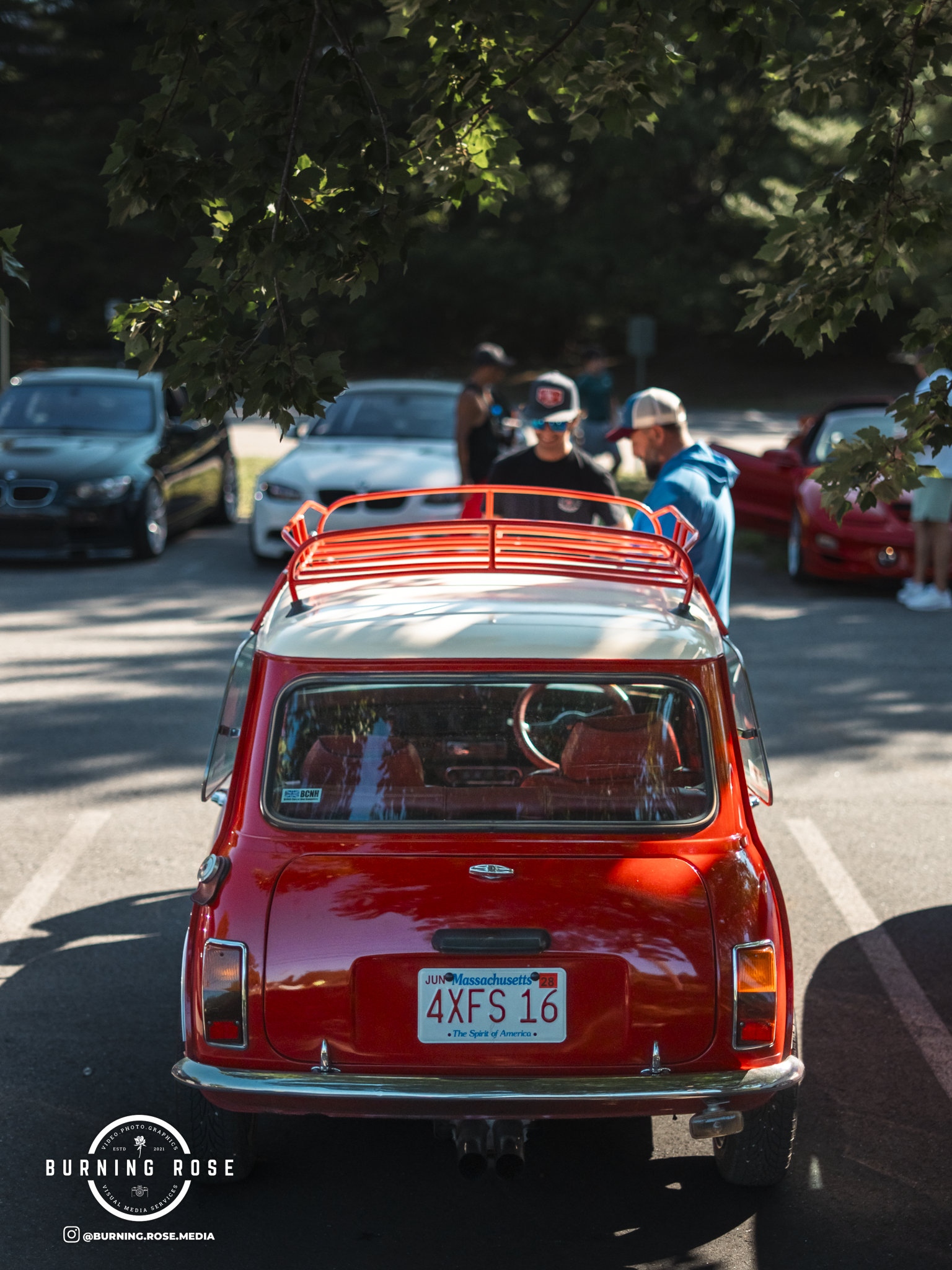 Red vintage car with Massachusetts license plate 'AXE 16' parked in a lot with other cars and people standing nearby. A tree branch with green leaves is visible at the top of the image.
