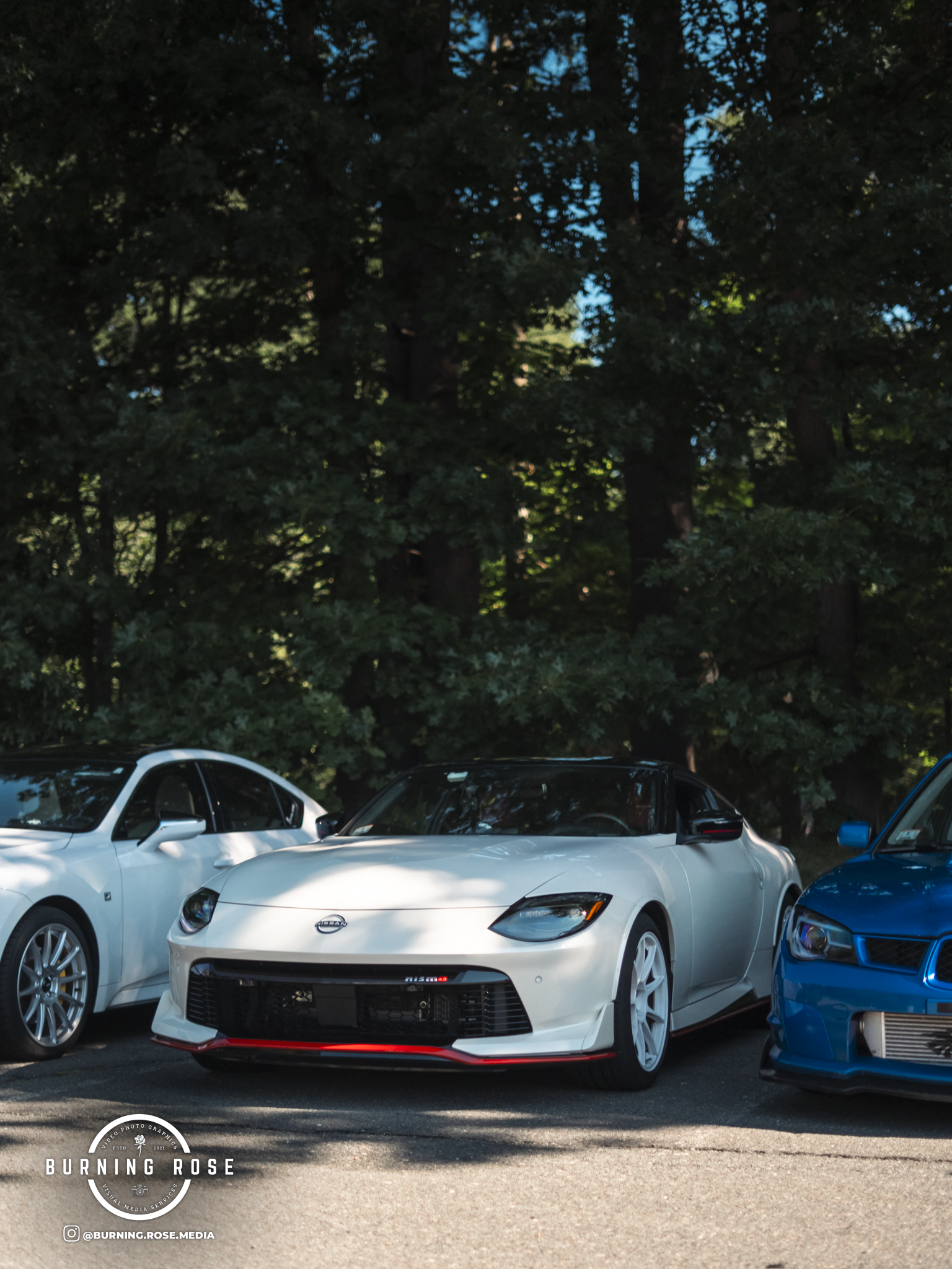 A white Nissan sports car parked between a blue car and a white car in a parking lot, with large trees in the background.