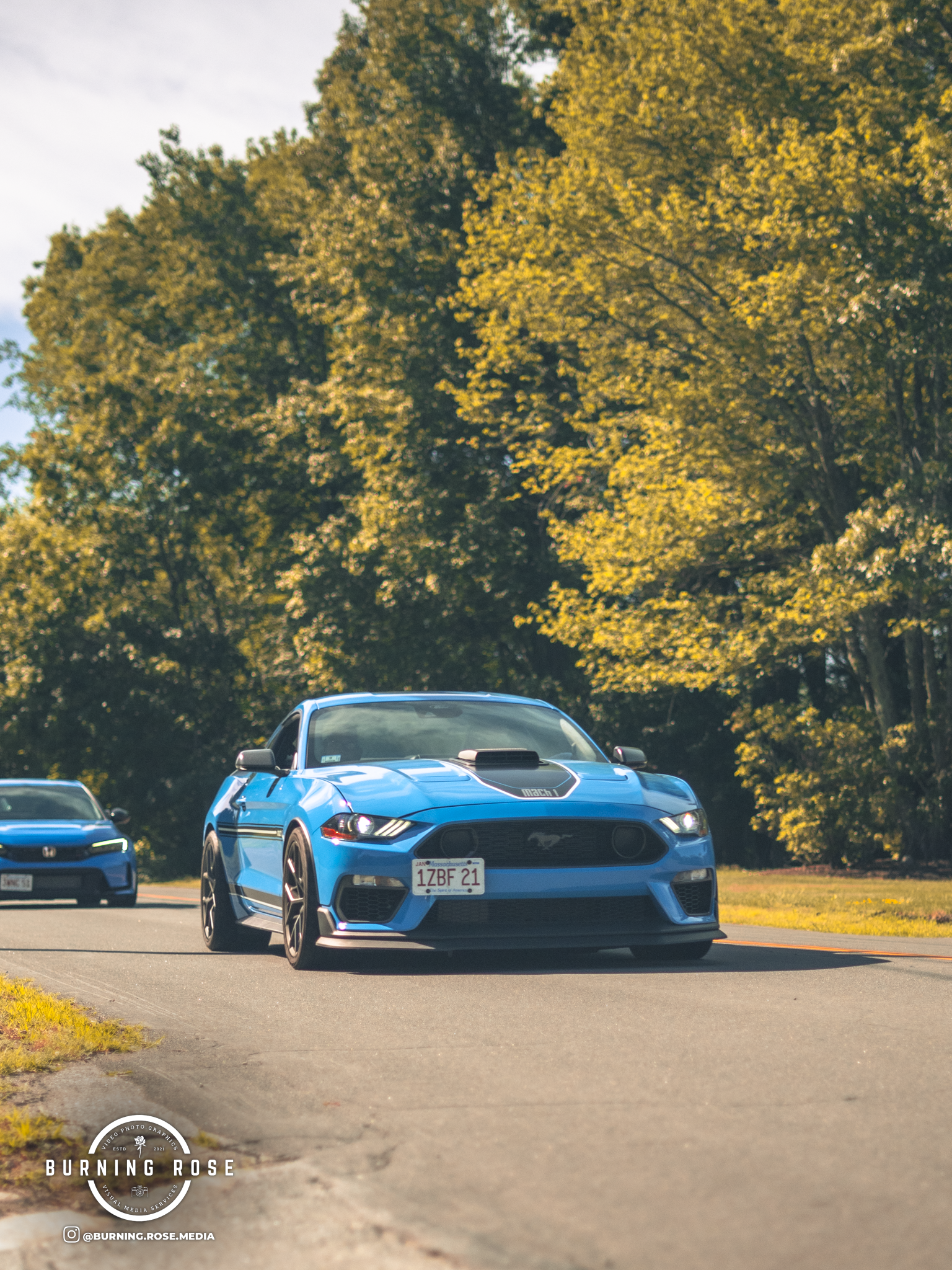 A bright blue Ford Mustang GT parked on a street with trees in the background foliage in fall colors. The car has an American license plate reading '1ZBF21', and there is another blue car partially visible on the left side.