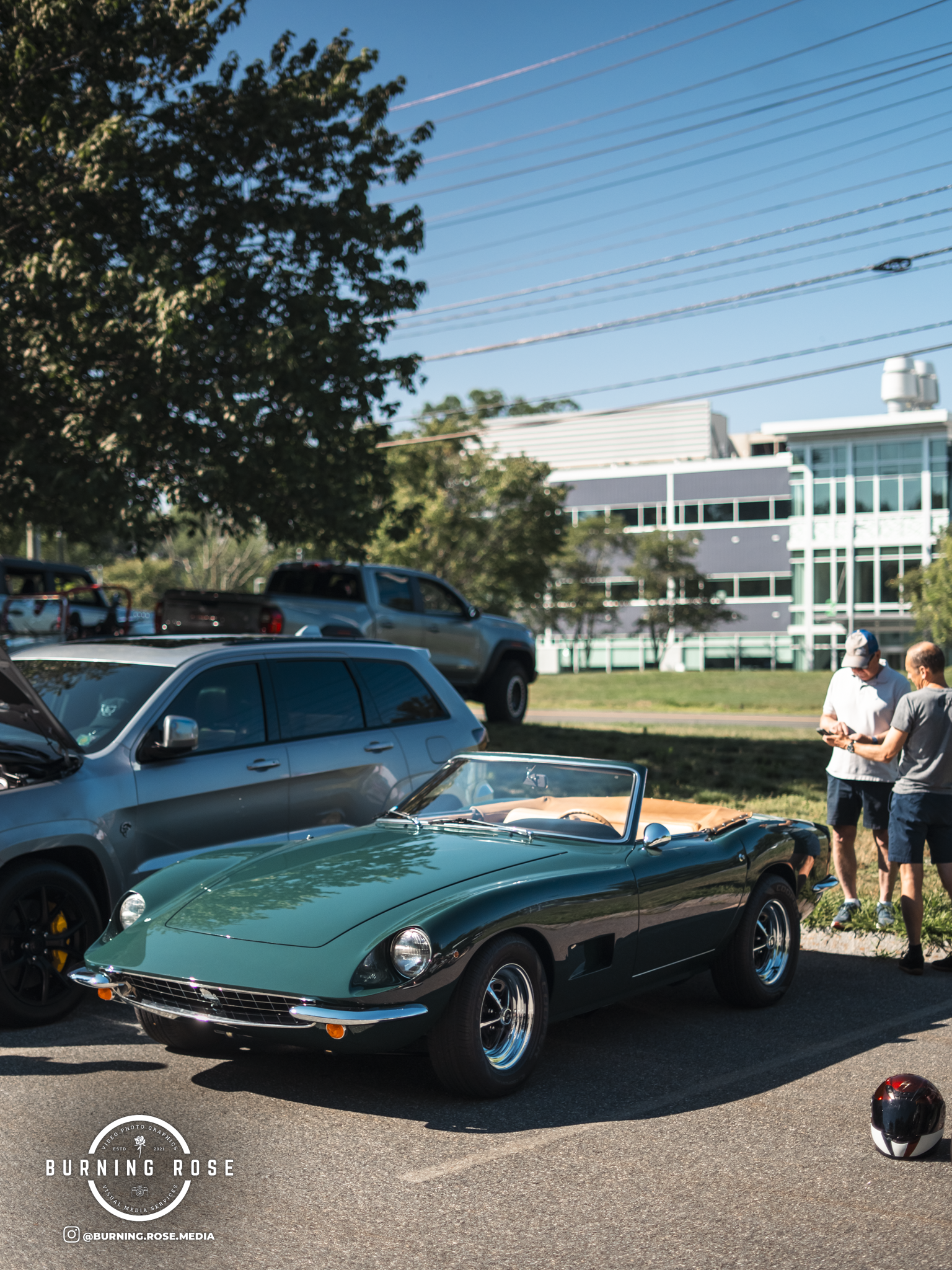 A sleek teal vintage convertible car parked outdoors at a car show, with two men standing nearby looking at their phones, and other cars and a modern building in the background.