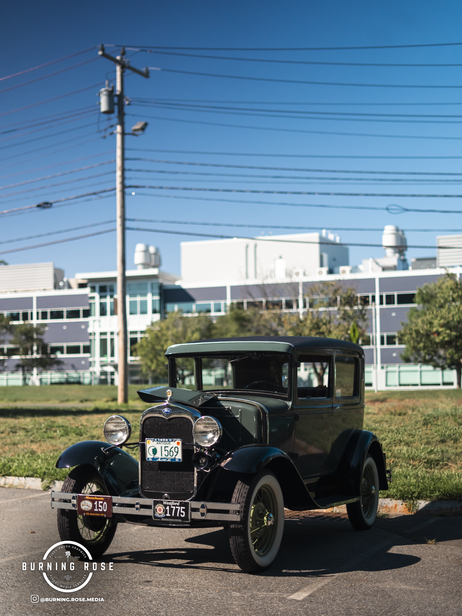A vintage black car parked on a street with a modern building, trees, power lines, and a clear blue sky in the background.