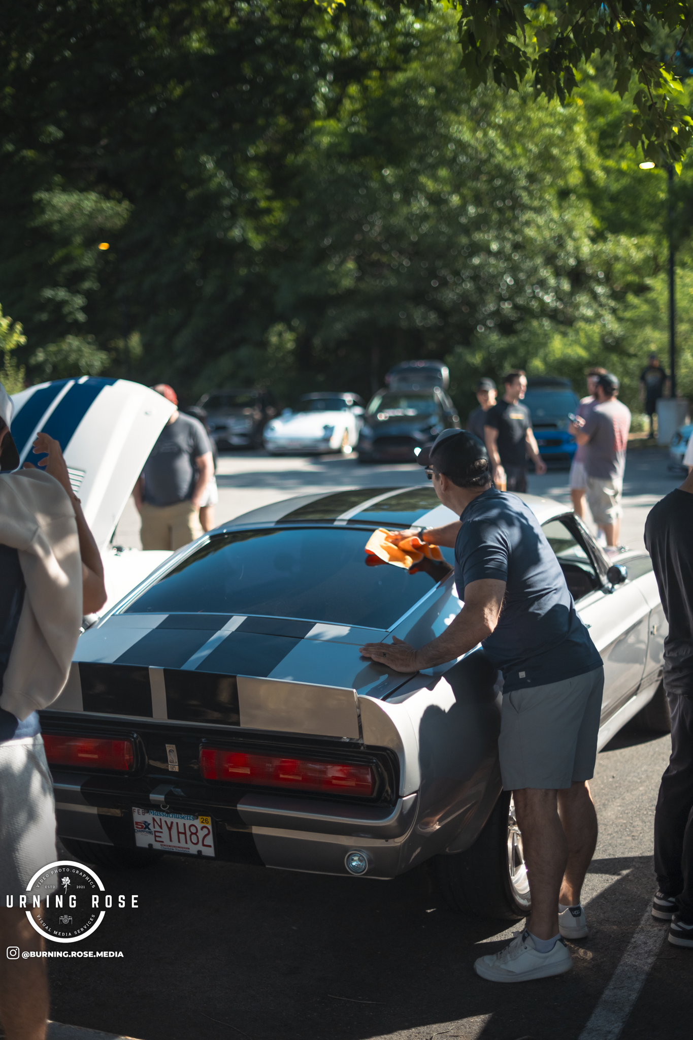 People gathered around a classic car with a silver and black striped design, outdoors in a parking lot with trees in the background, during what appears to be a car meet or showcase.