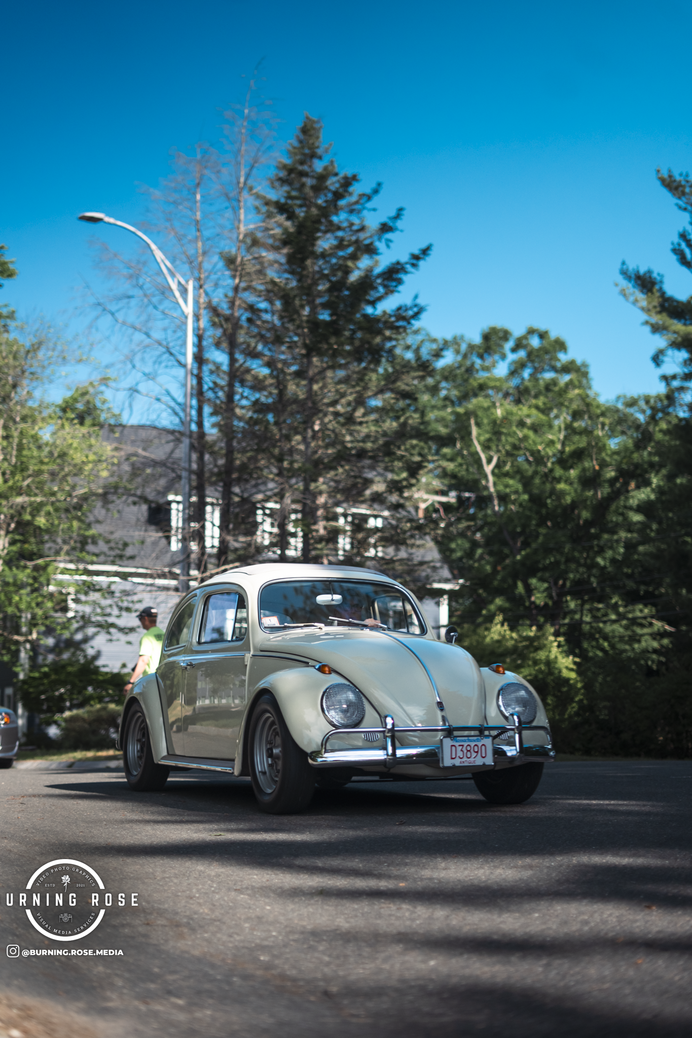 A vintage beige Volkswagen Beetle car parked on a street with trees and a house in the background. There is a person wearing a hat and neon green shirt walking beside it, and a streetlamp is visible in the scene.