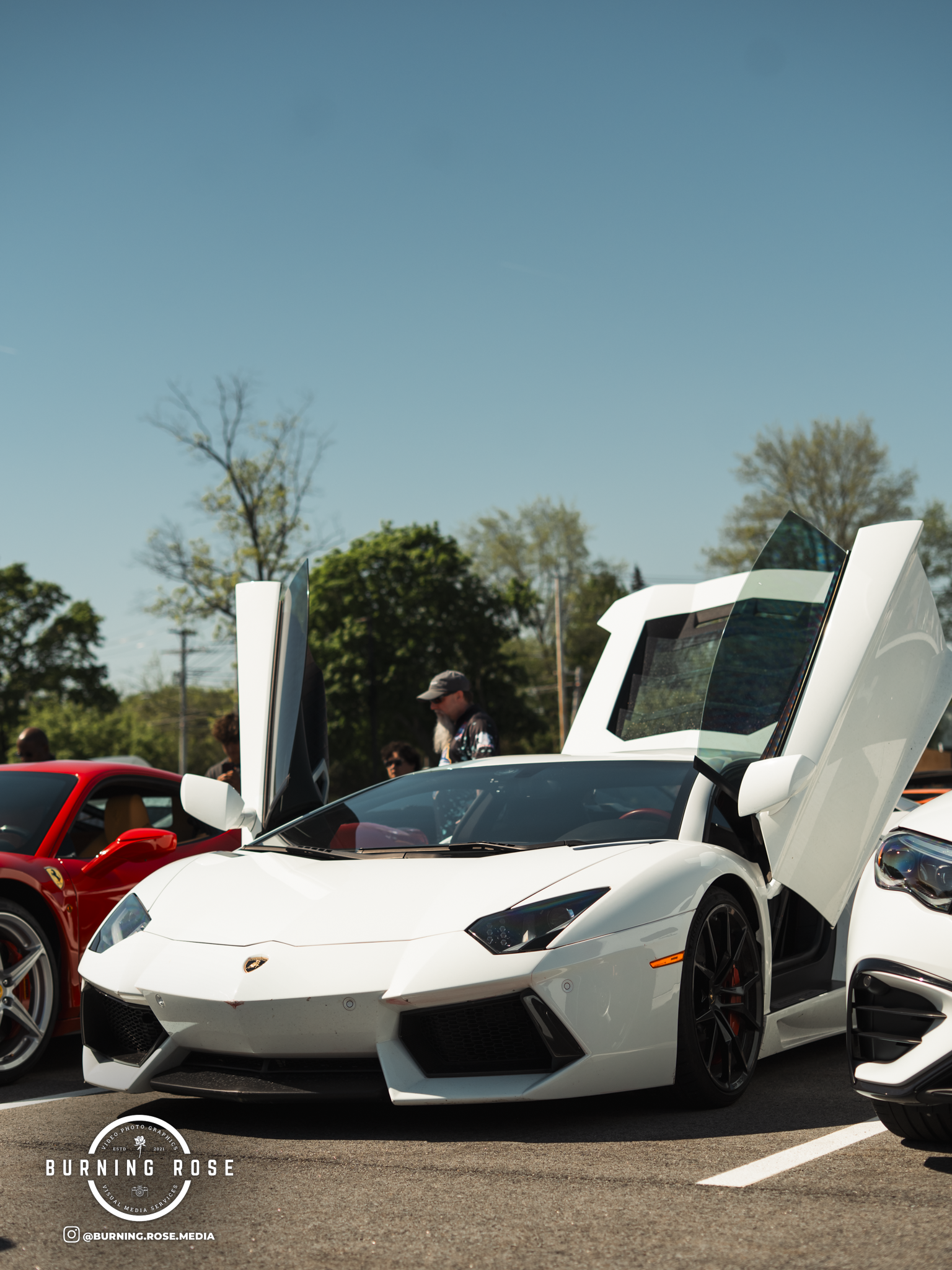 A white Lamborghini sports car with its door open, parked in a lot at a car show with other vehicles and people in the background.