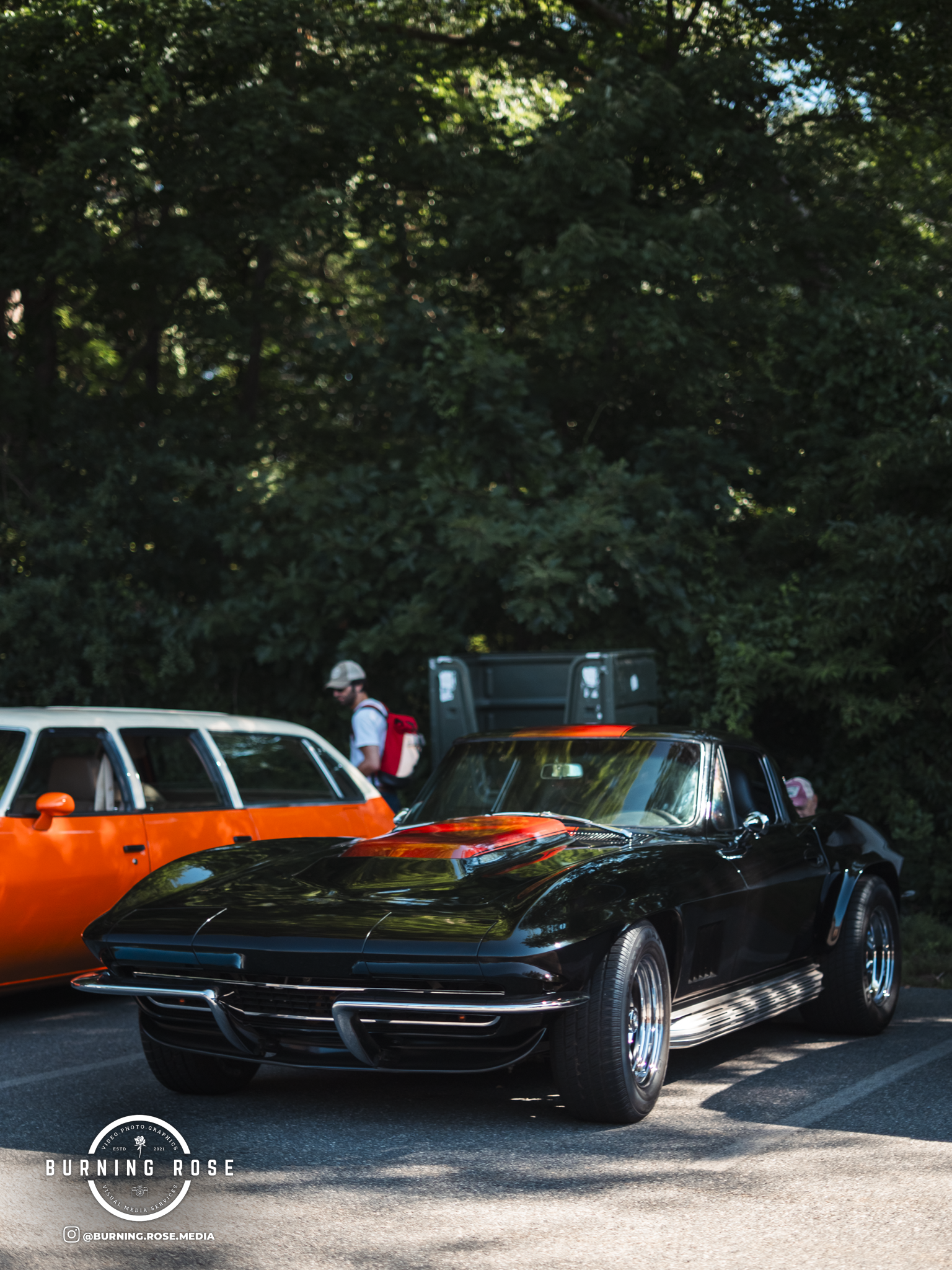 A black vintage car parked in front of a large green tree, with a person walking nearby and other classic cars partially visible in the background.