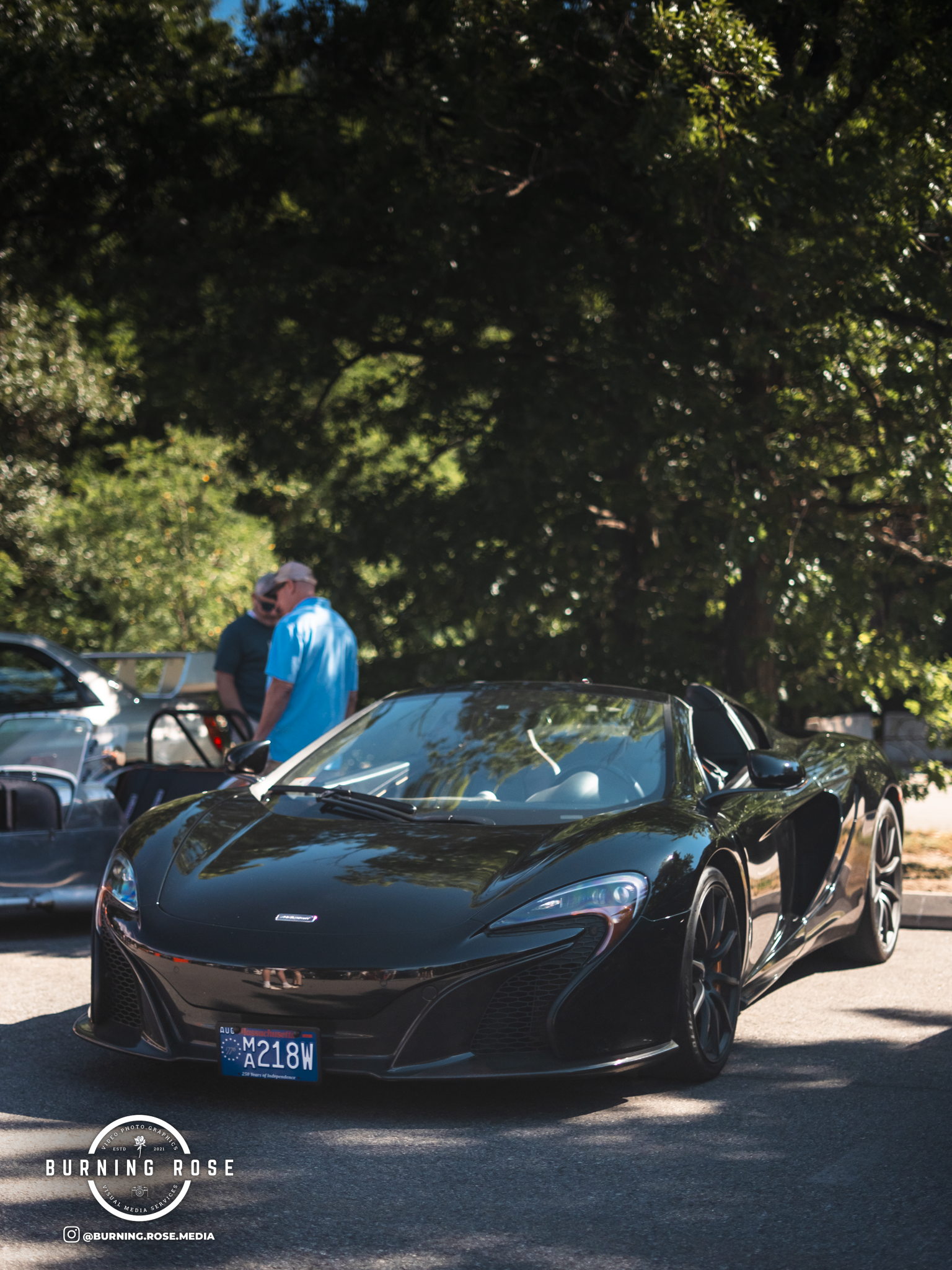 A black sports car parked outdoors under trees, with two men standing beside it, engaging in conversation.