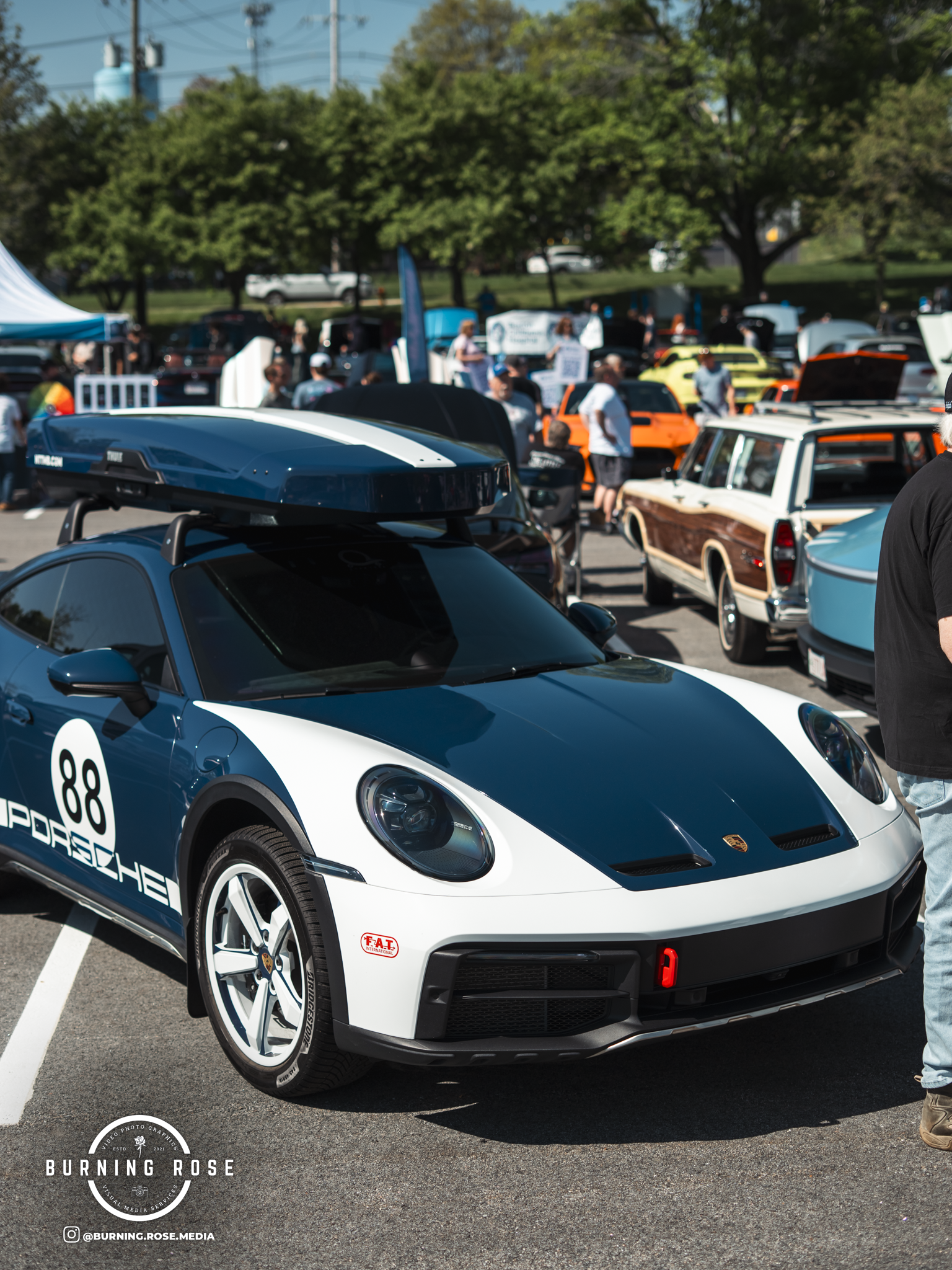 A blue and white Porsche sports car with the number 89 on the side is parked in a crowded outdoor car show. The background shows other classic and modern cars, people, and trees.