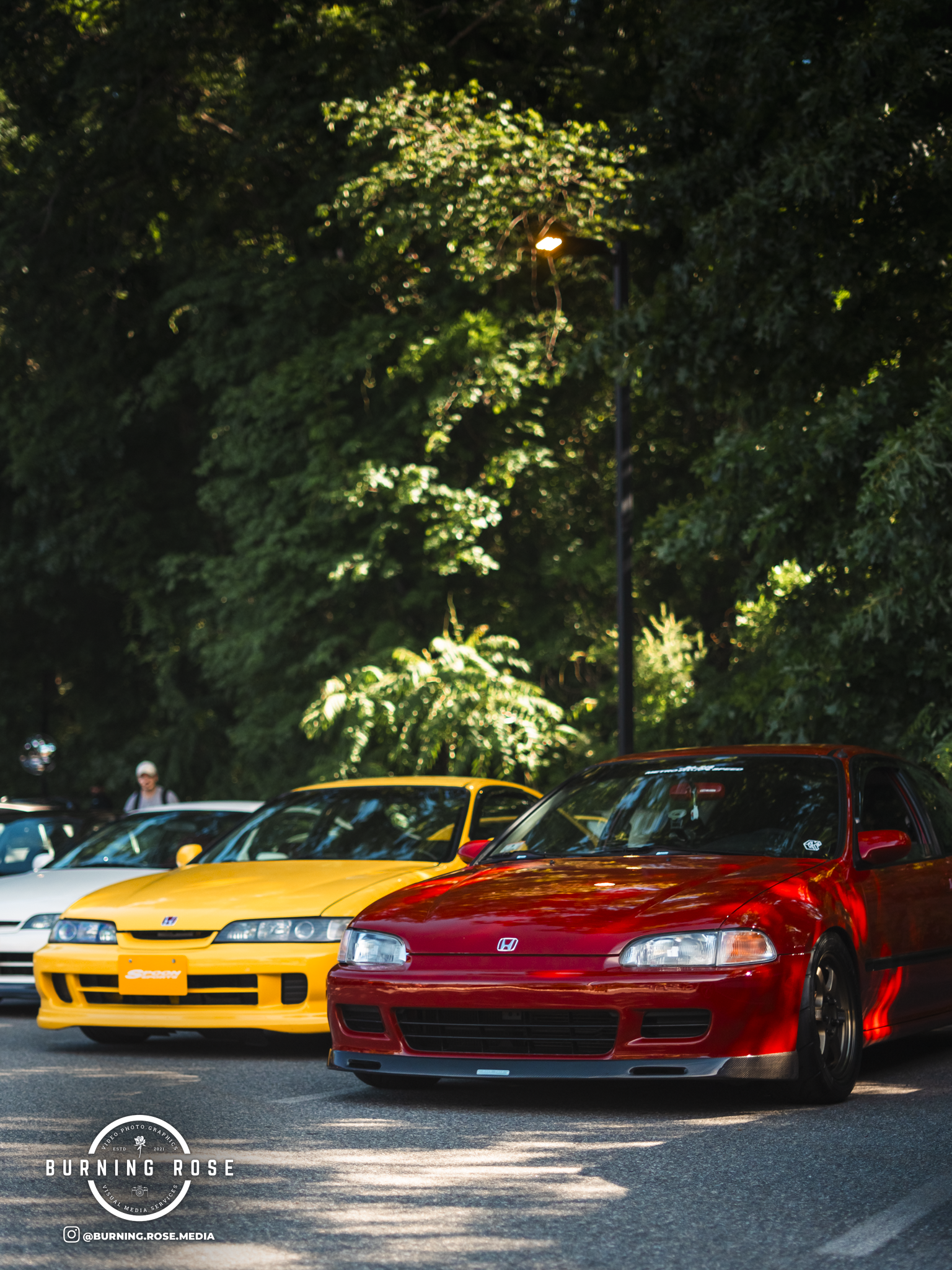 Red Honda car parked in front of a yellow car with the parking lot and green trees in the background.