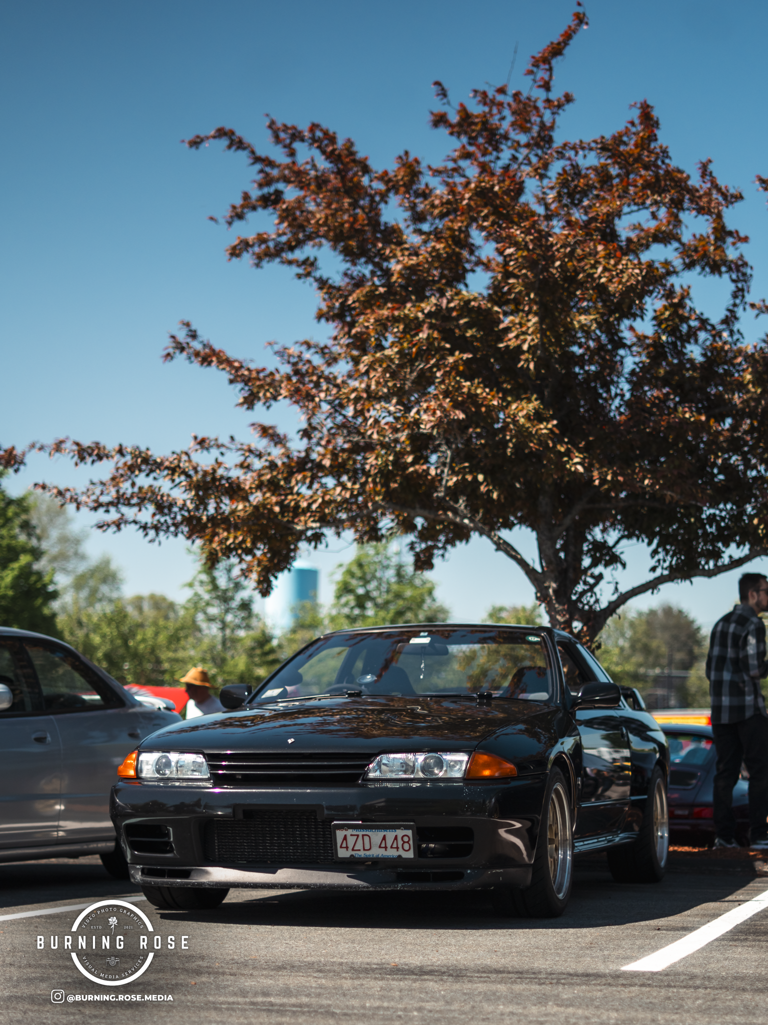 Close-up of a black sports car parked in a parking lot with a tree and other vehicles in the background, under a clear blue sky.