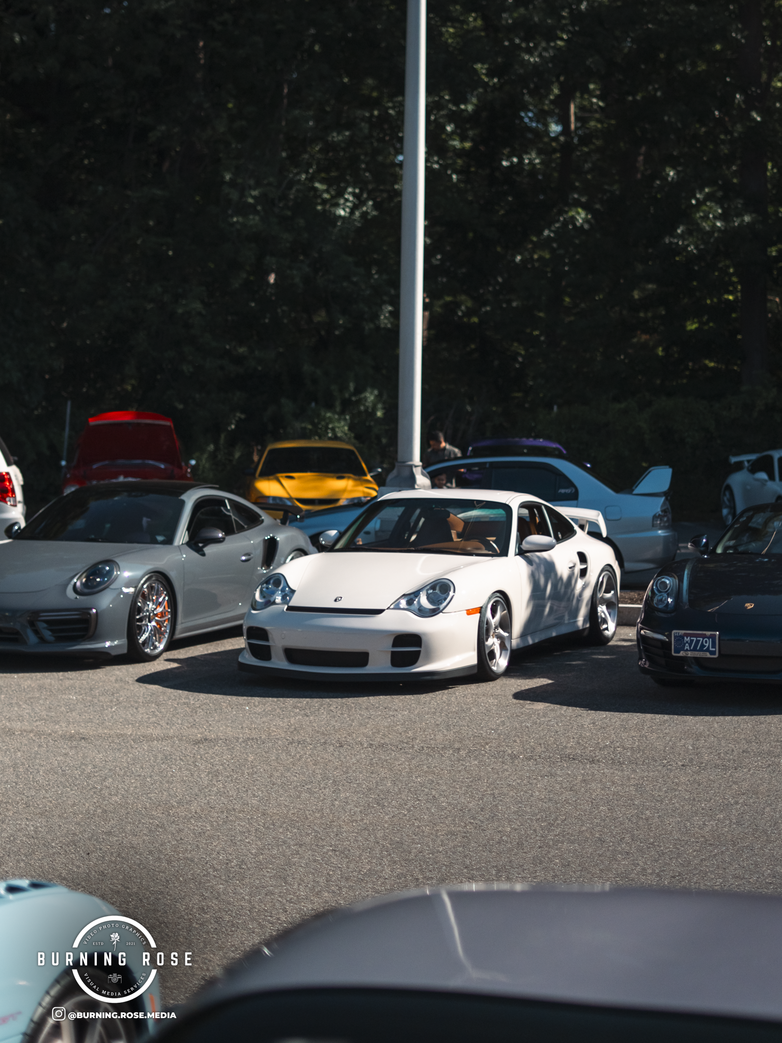 A parking lot filled with various colored sports cars, including a prominent white Porsche in the foreground, under a clear sky with dark trees in the background.