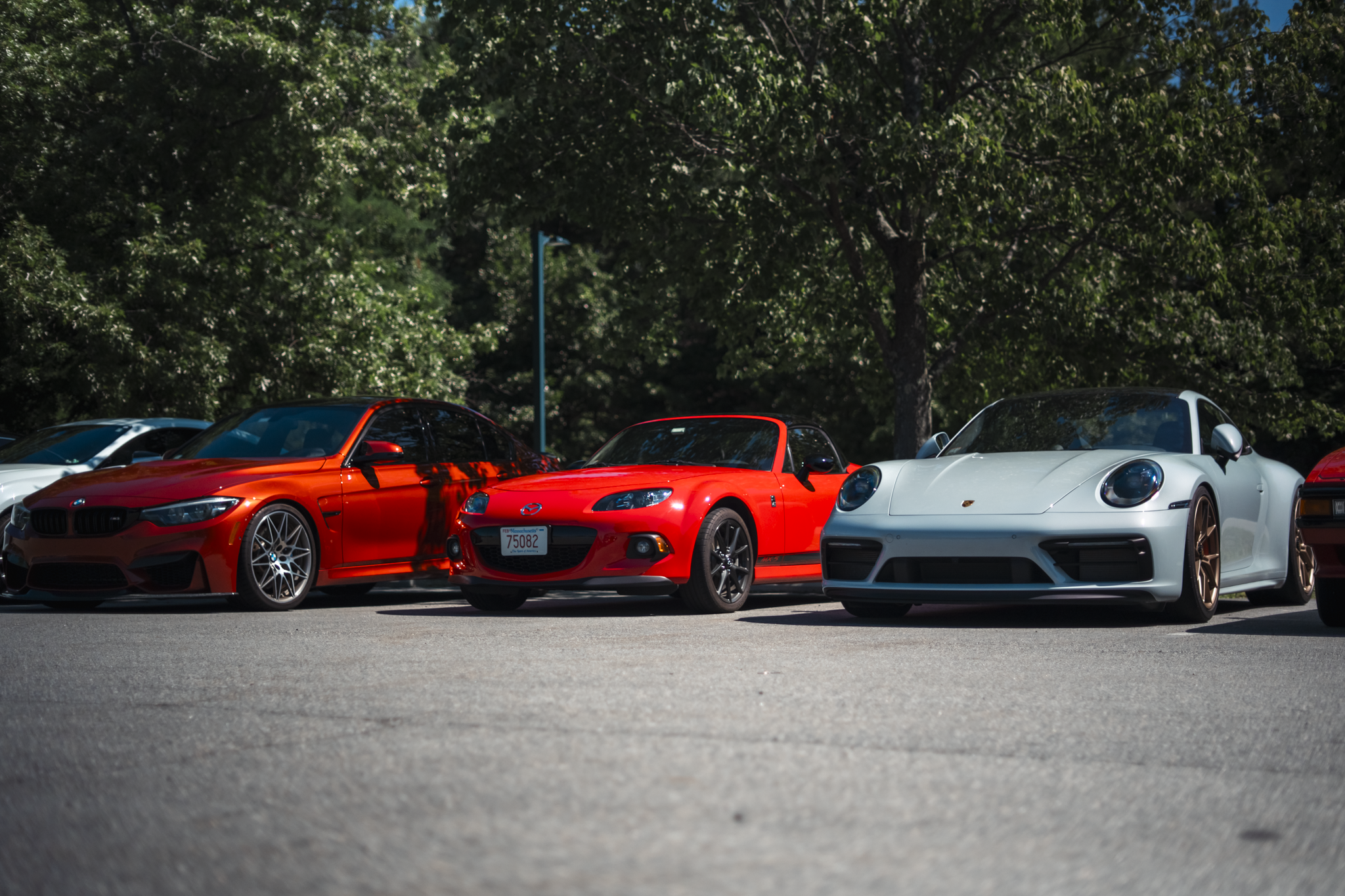 A row of sports cars parked outdoors, including a red Mazda MX-5 Miata, a white Porsche, and a black BMW with a sunset reflected on their surfaces, with trees and a blue sky in the background.