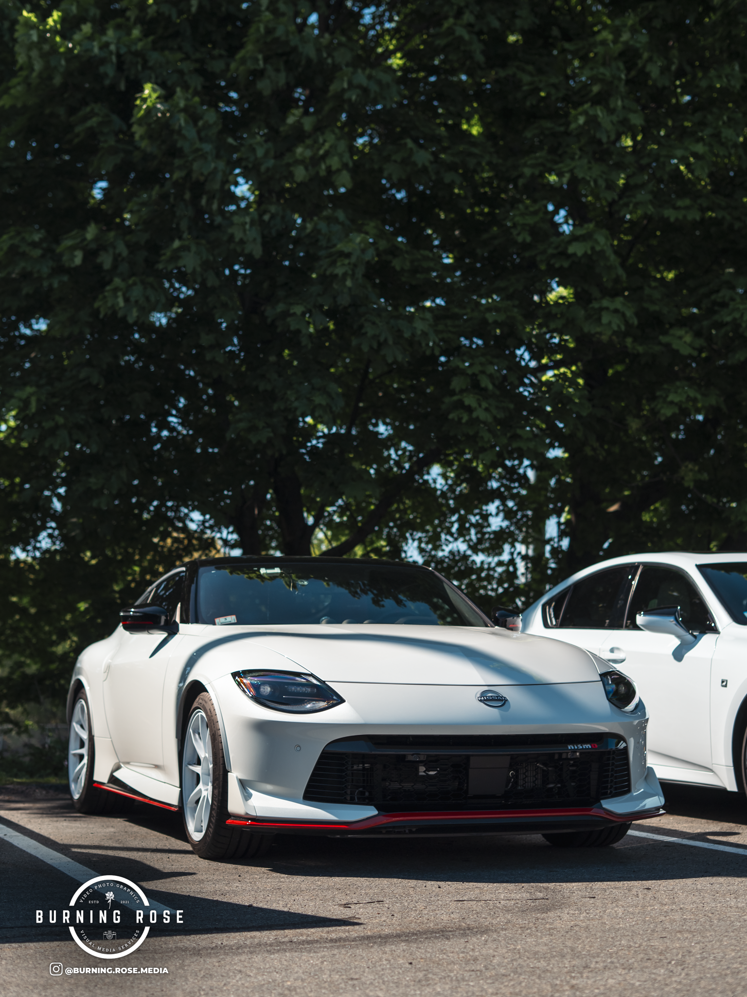 A white sports car parked in an outdoor parking lot with a second white vehicle visible in the background, surrounded by green trees and blue sky.
