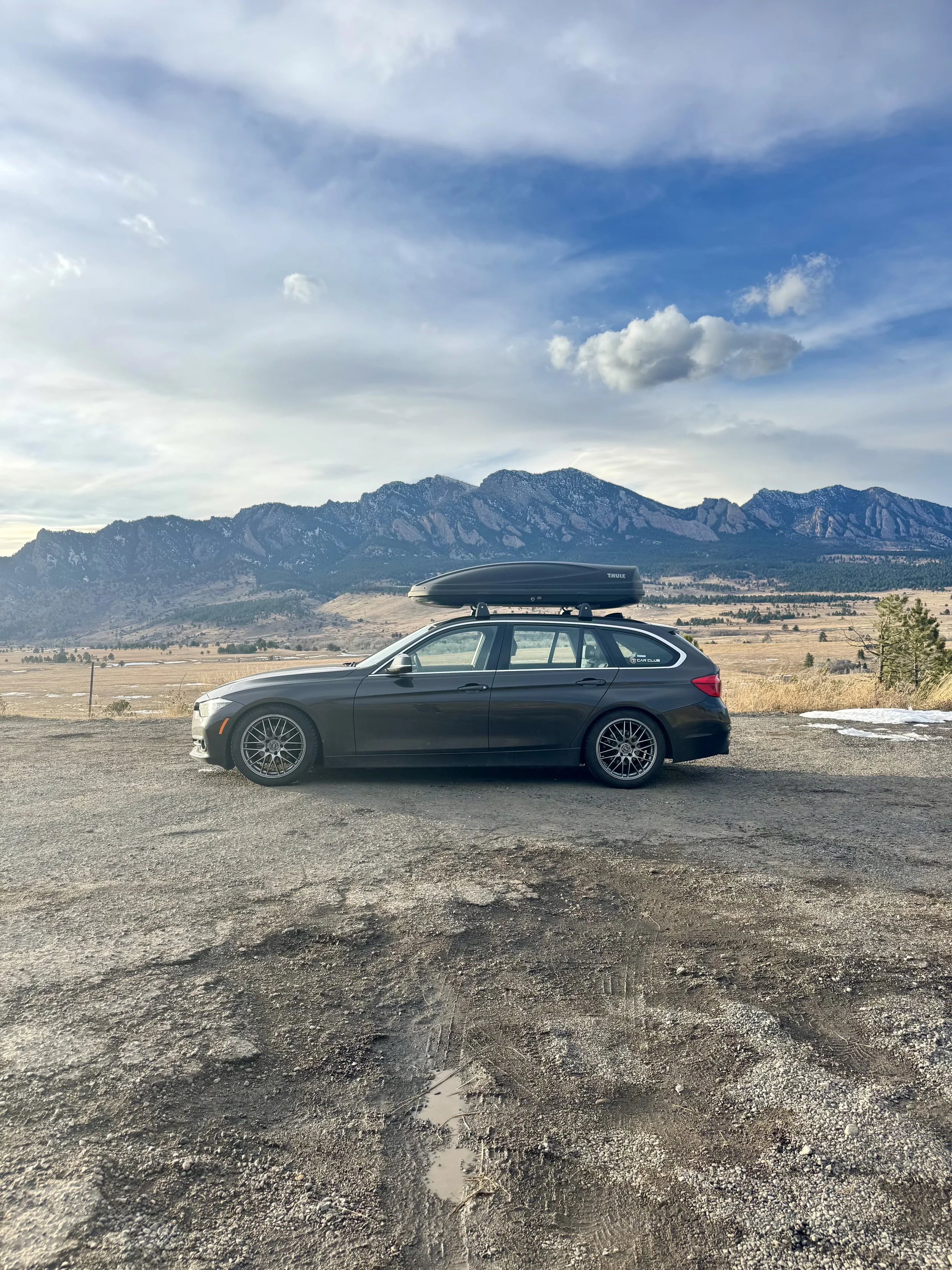 A black BMW station wagon parked on a driveway beside a light gray building, with green grass and trees in the background under a partly cloudy sky.