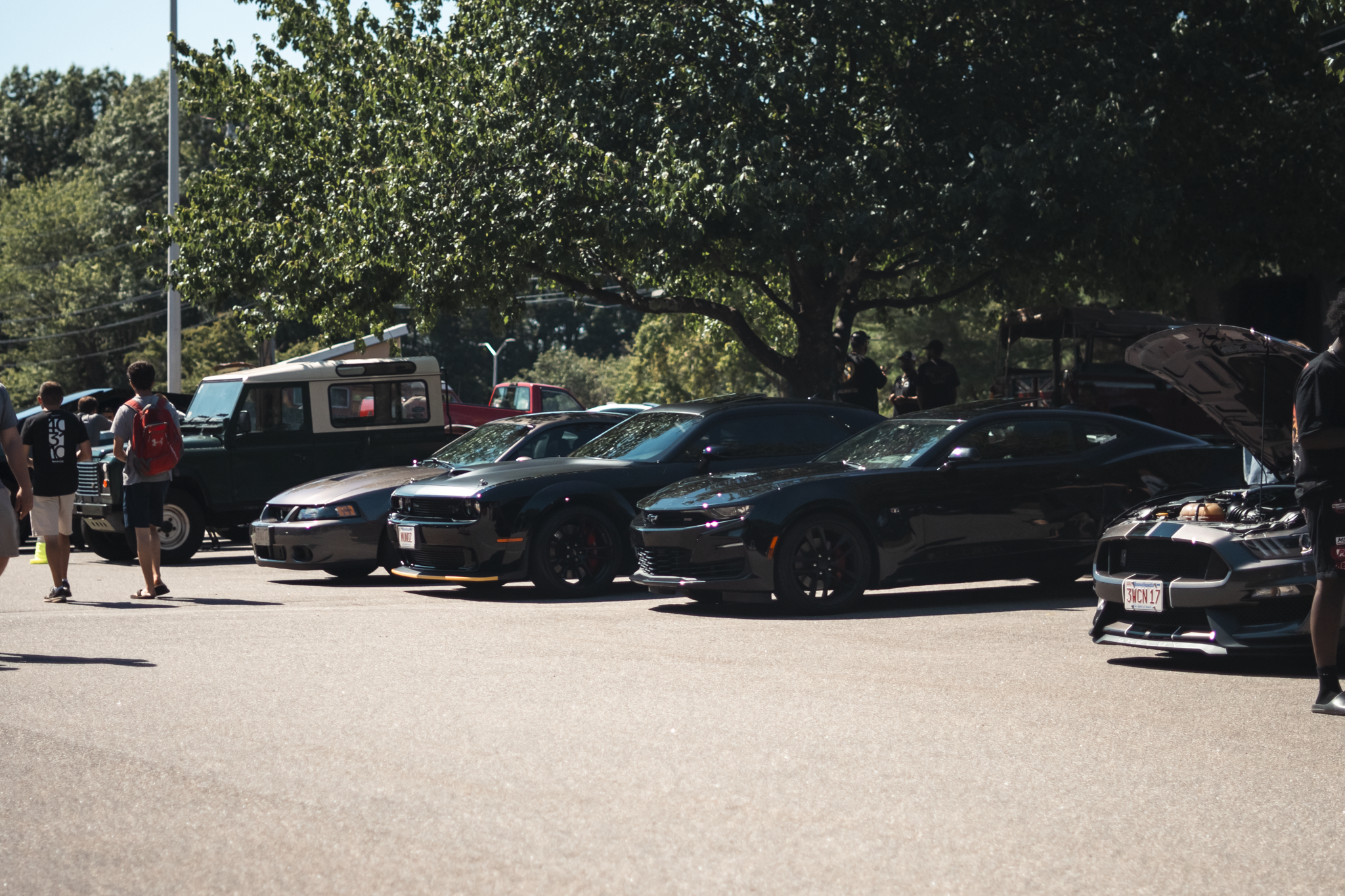A parking lot filled with several parked cars including a black sports car and a gray sports car, with a group of people walking nearby and trees in the background on a sunny day.