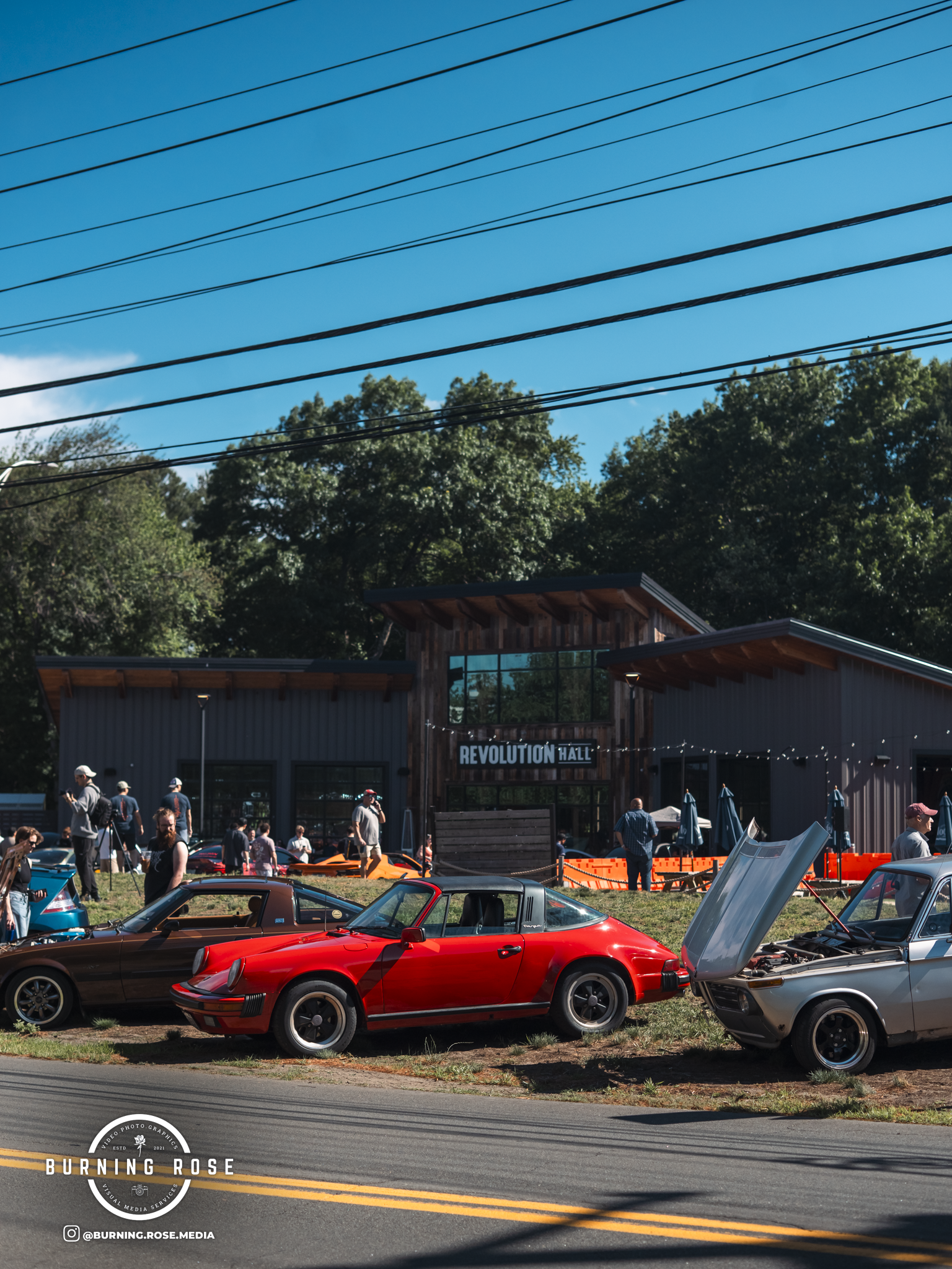 Classic and sports cars on display at a car show, with a building labeled 'Revolution Hall' in the background, under a clear blue sky and trees.