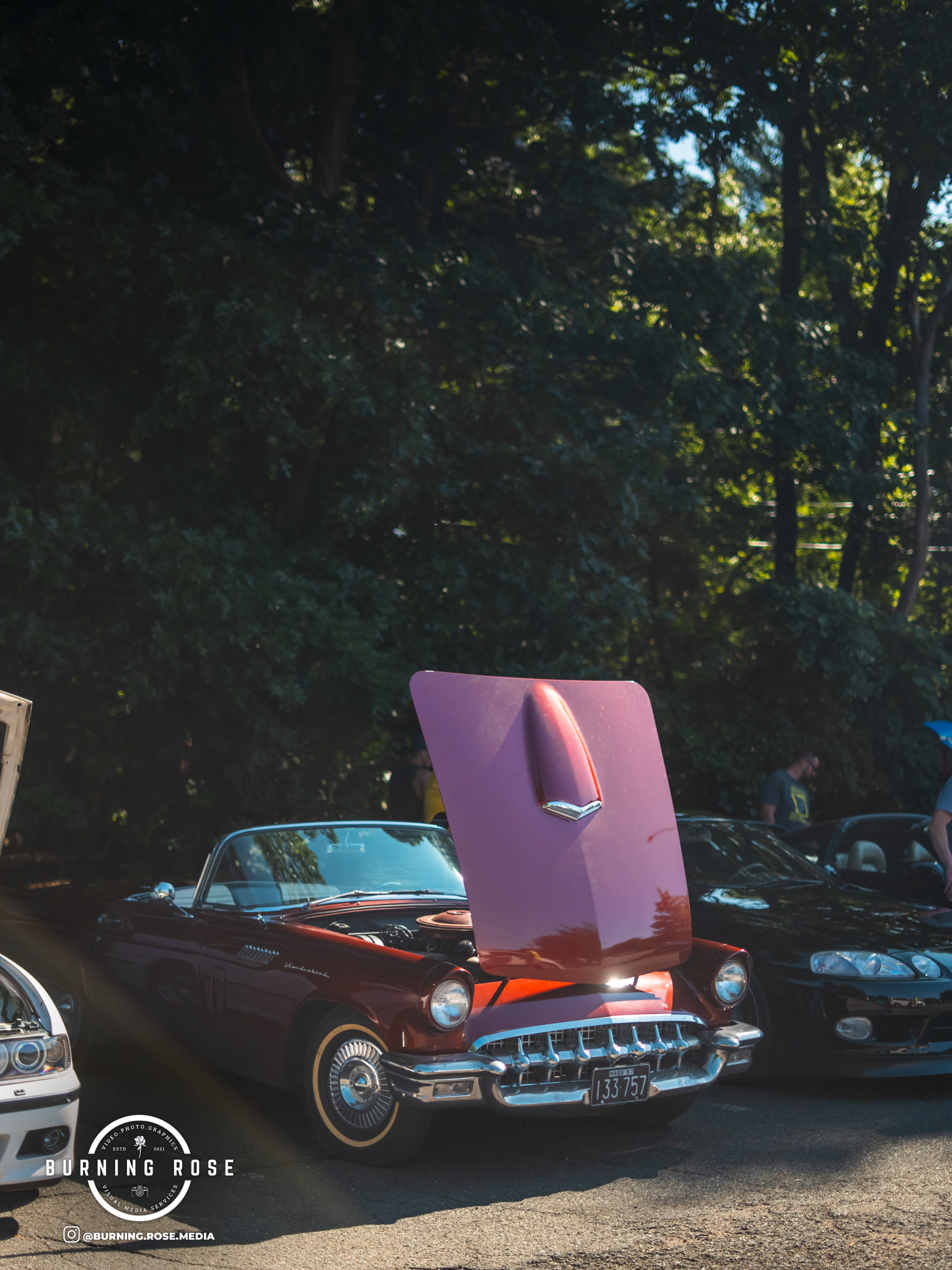 A classic red Ford Mustang with its hood open at a car show, surrounded by other vintage cars and parking lot attendees.
