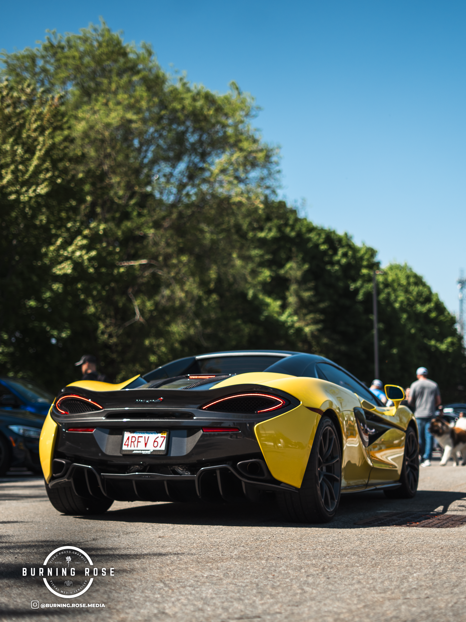 A yellow sports car with a black rear diffuser and distinctive taillights parked outdoors, with trees and people in the background, under a clear blue sky.