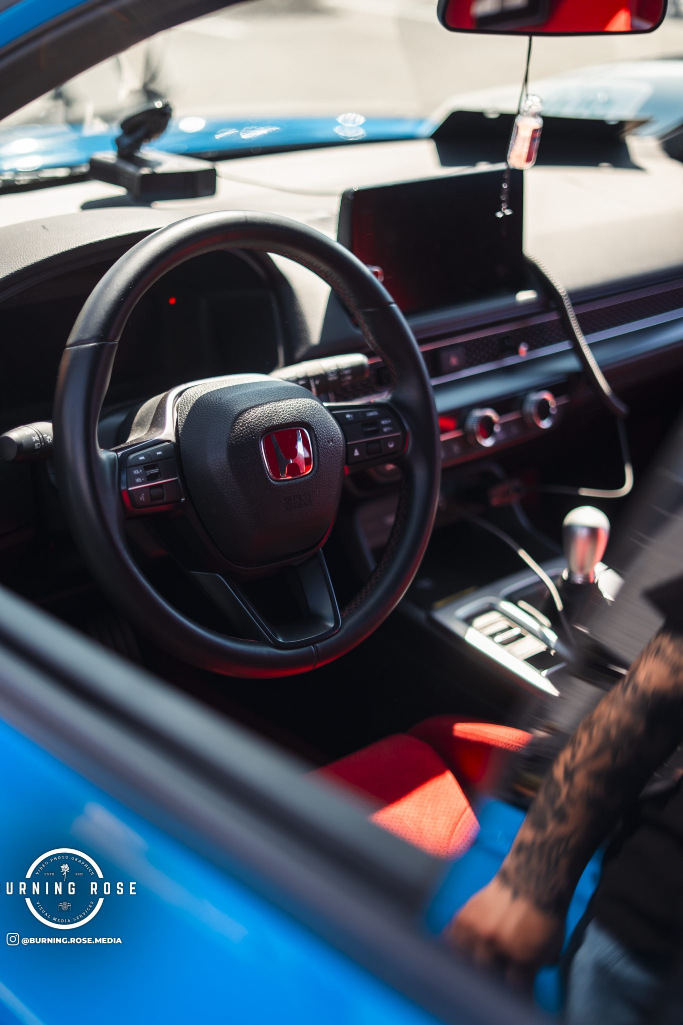 The interior of a car showing the steering wheel with Honda logo, dashboard, center console, and a red seat or cushion on the driver's seat. Sunlight illuminates parts of the interior through the windows.