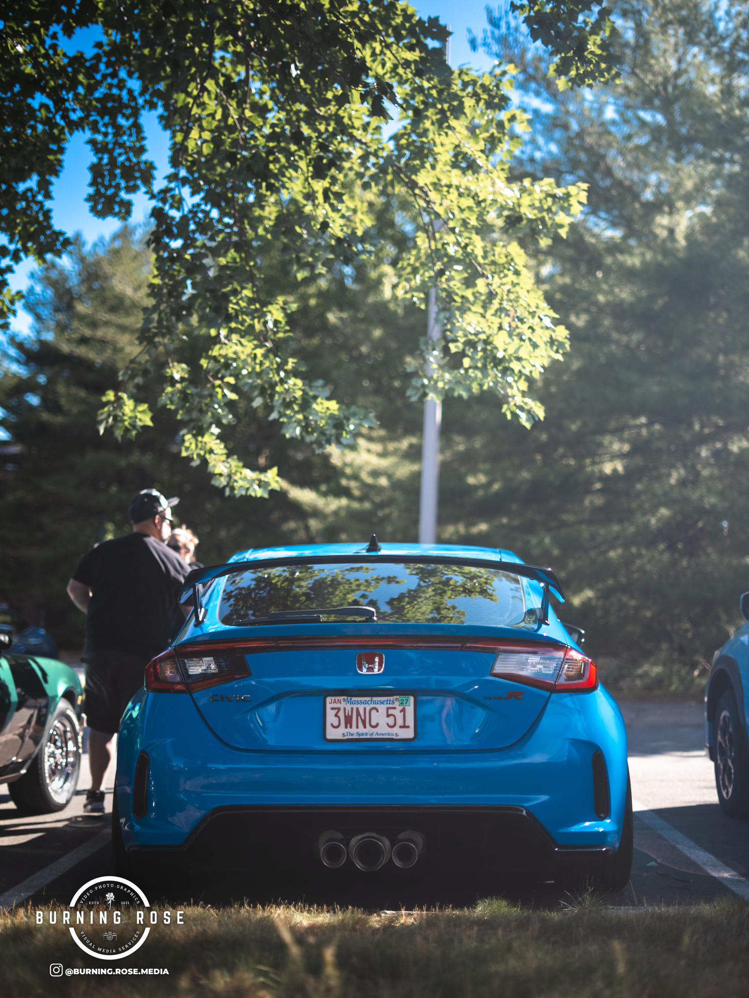 Rear view of a bright blue Honda Civic Type R parked outdoors under a tree, reflecting the green leaves and blue sky on its rear windshield, with a person and a green car visible on the left side.