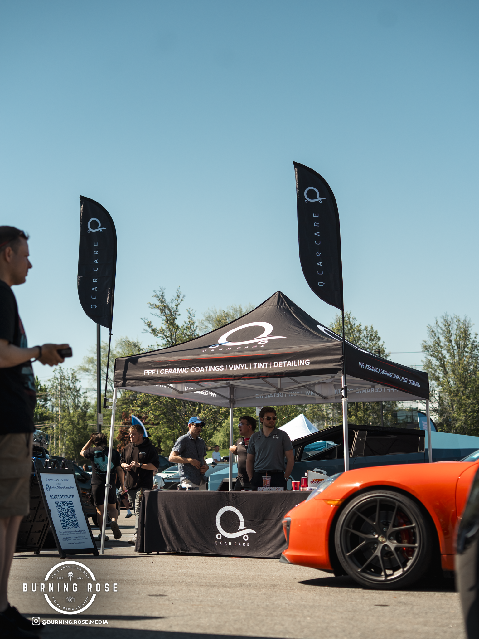 At an outdoor auto event, a bright orange sports car is visible in the foreground. Behind it, a canopy tent marked 'QCARCARE' advertises car detailing services like PPF, ceramic coatings, vinyl, tint, and detailing. Several people are gathered around