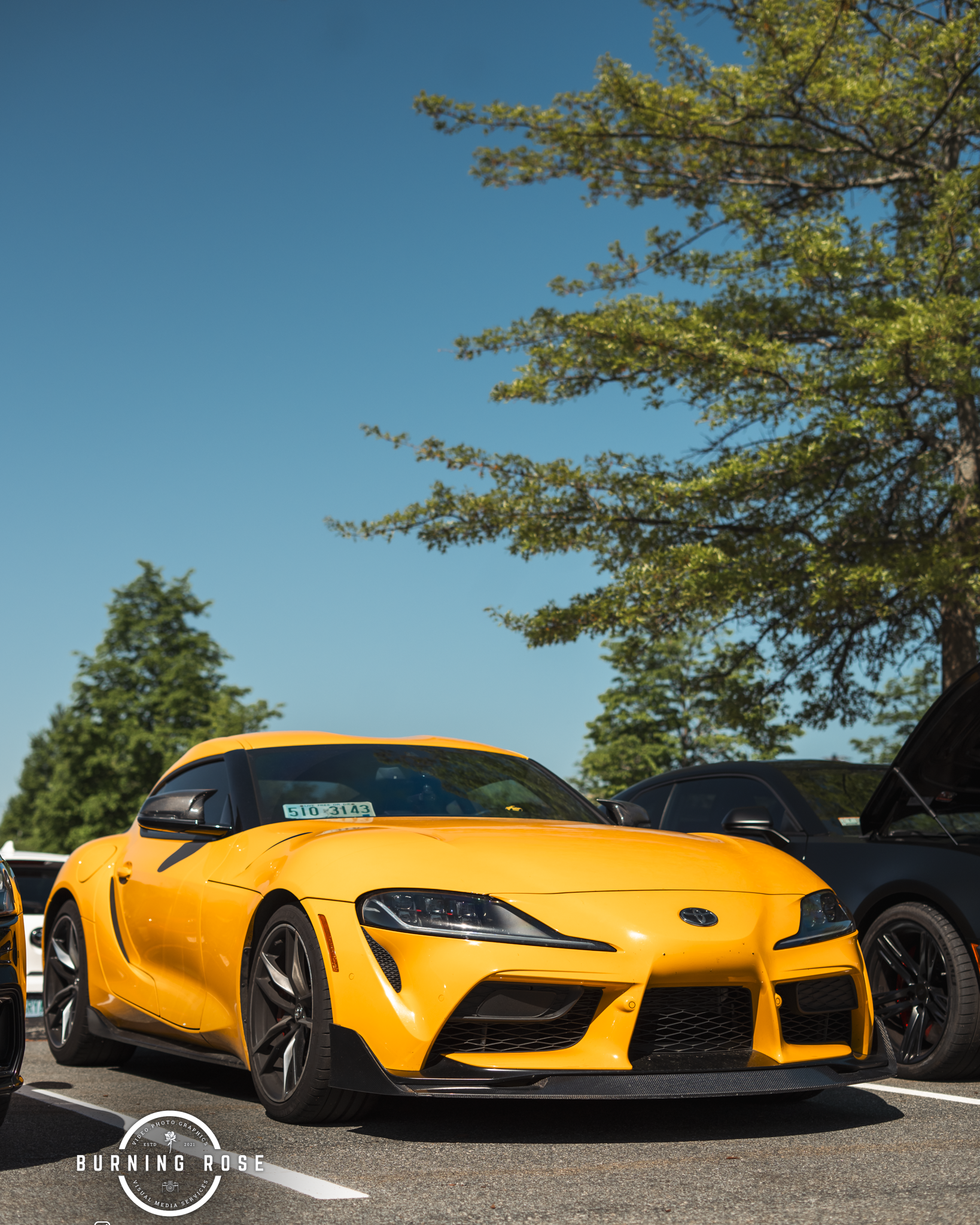 A yellow sports car parked in a parking lot under a clear blue sky with trees in the background.