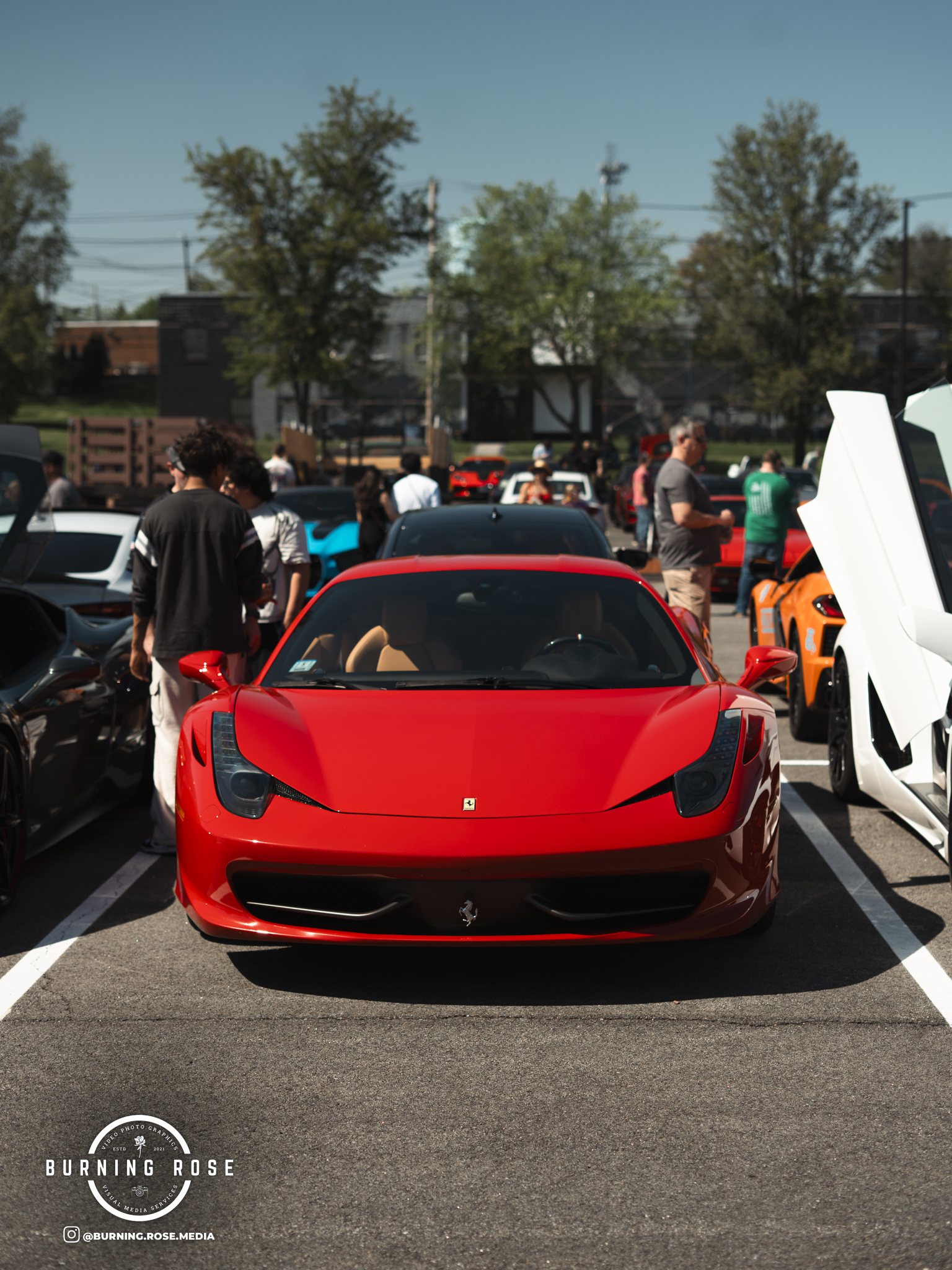 A red Ferrari sports car parked in a lot with people and other cars, including a white and an orange car, visible in the background under a clear blue sky.