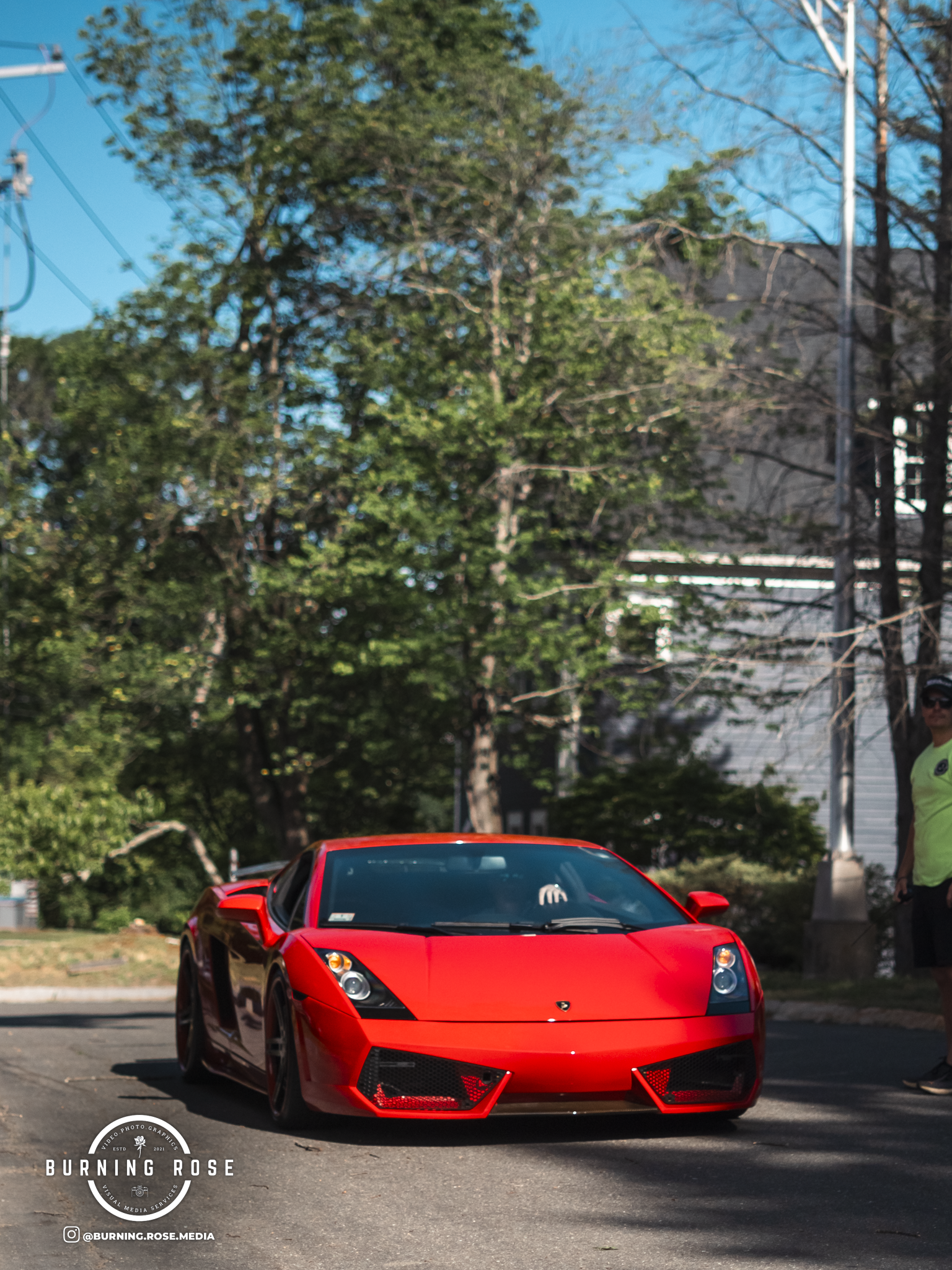 A red Lamborghini parked on a street with trees and a man wearing sunglasses and a yellow shirt nearby.
