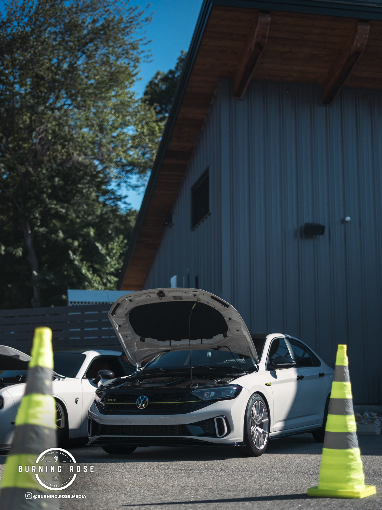 A white Volkswagen GTI with hood open at a car meet, surrounded by green traffic cones, in front of a blue building and trees.