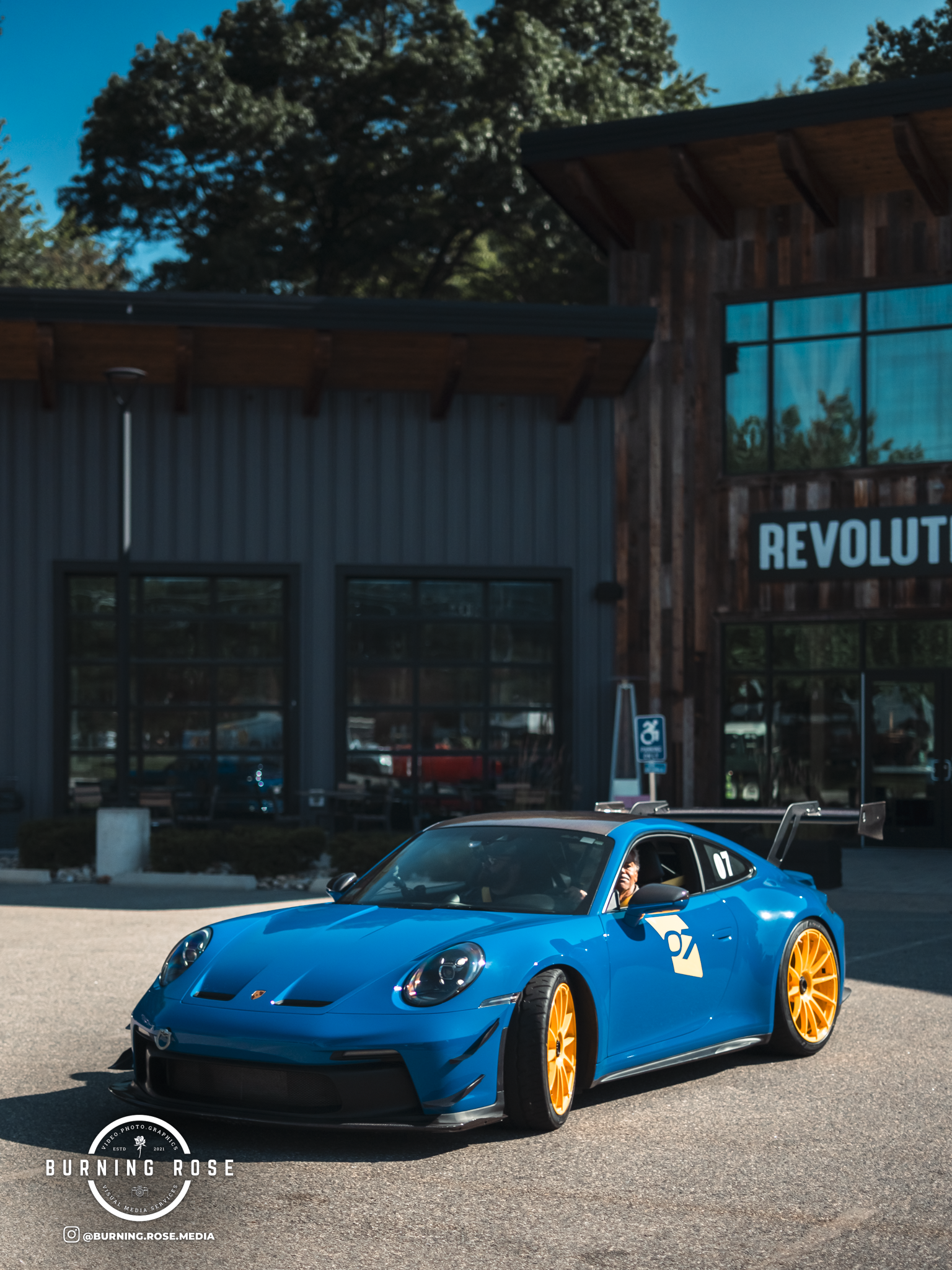 A blue sports car with orange wheels driving on a parking lot in front of a building with large windows and a sign that says 'Revolution'.