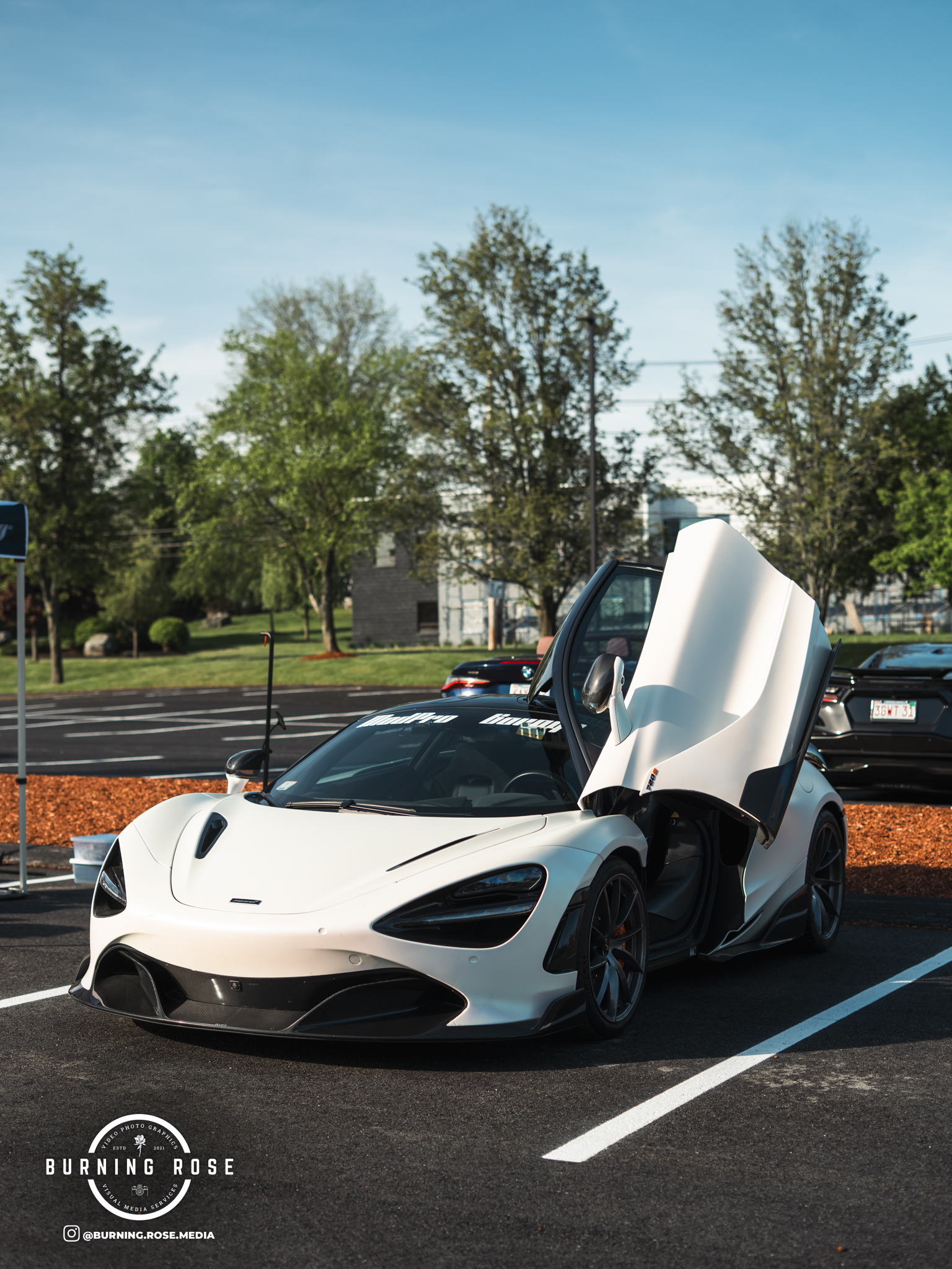 White sports car with open scissor doors parked in a parking lot with trees and a building in the background during daytime.