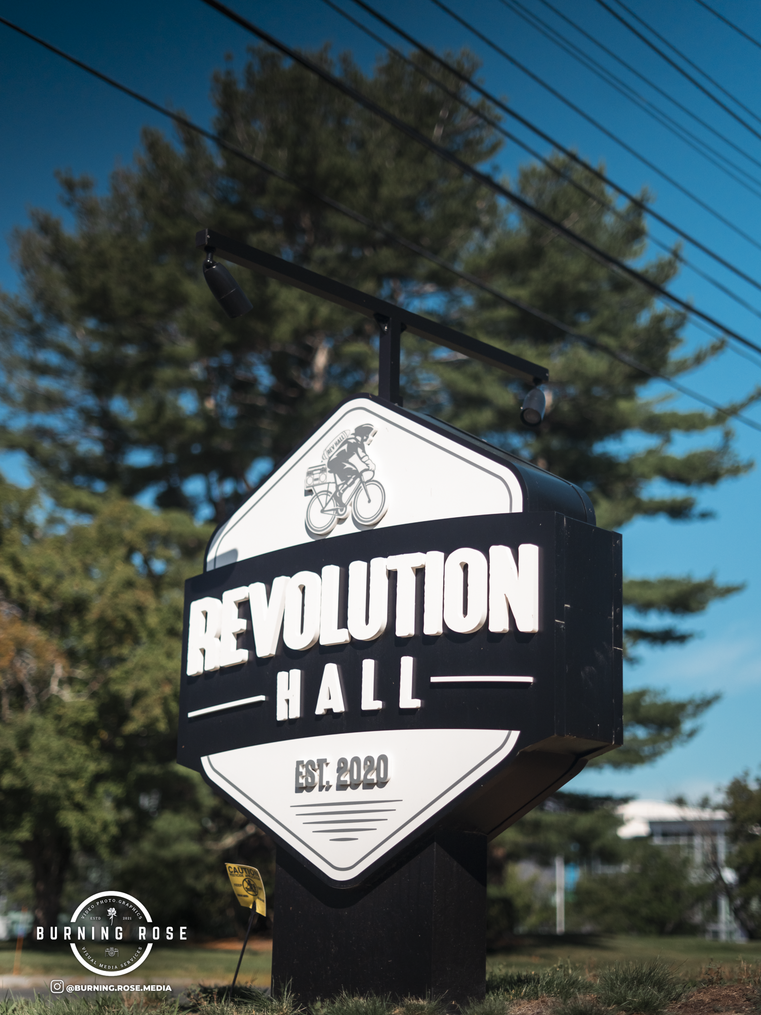 A black and white signboard with the text "REVOLUTION HALL" and an illustration of a person riding a bicycle, mounted on a pole with outdoor background including trees and power lines. The sign also states "EST. 2020" at the bottom.