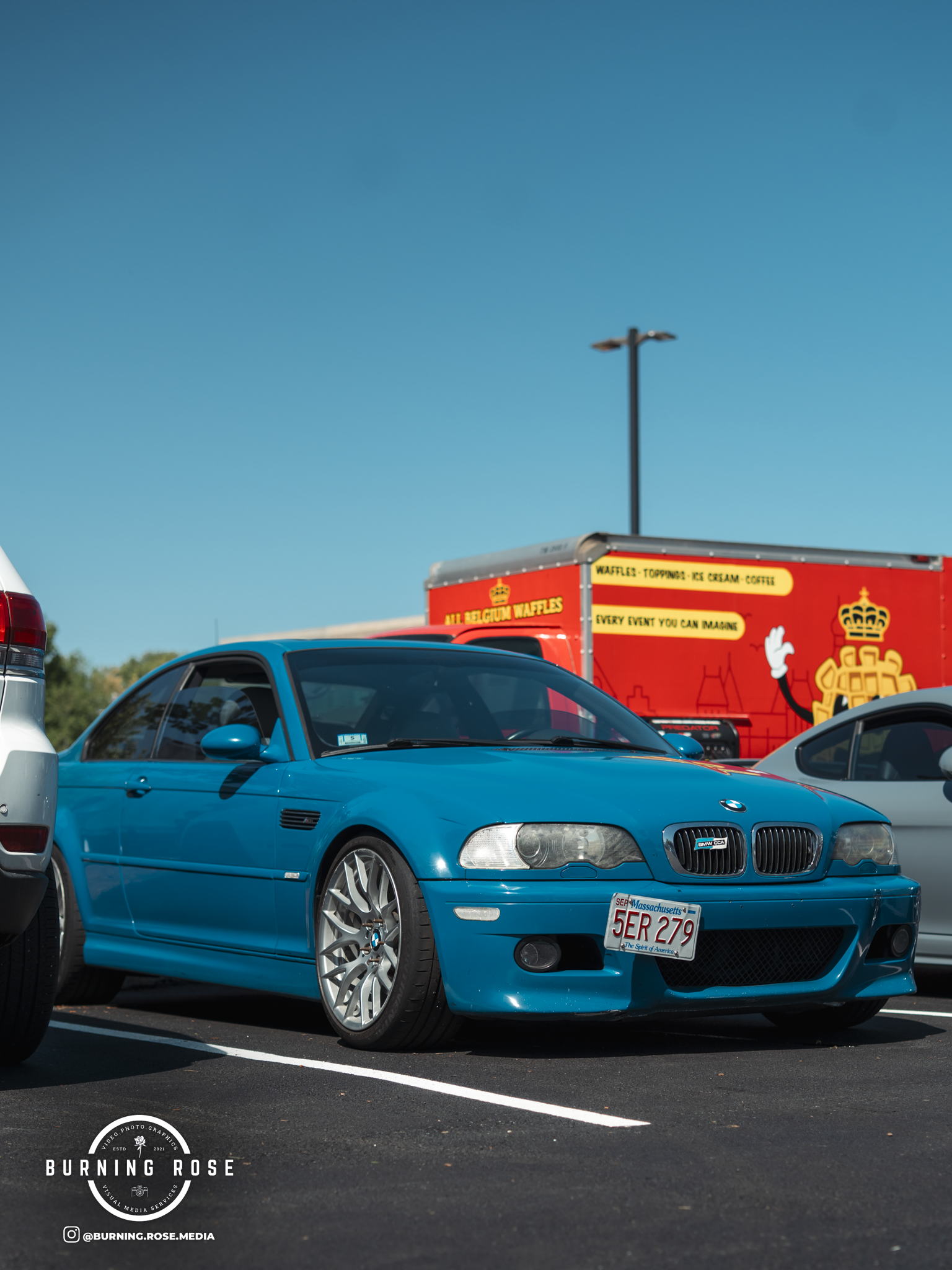 Blue BMW convertible parked in a parking lot, with a red food truck and other cars in the background under a clear blue sky.