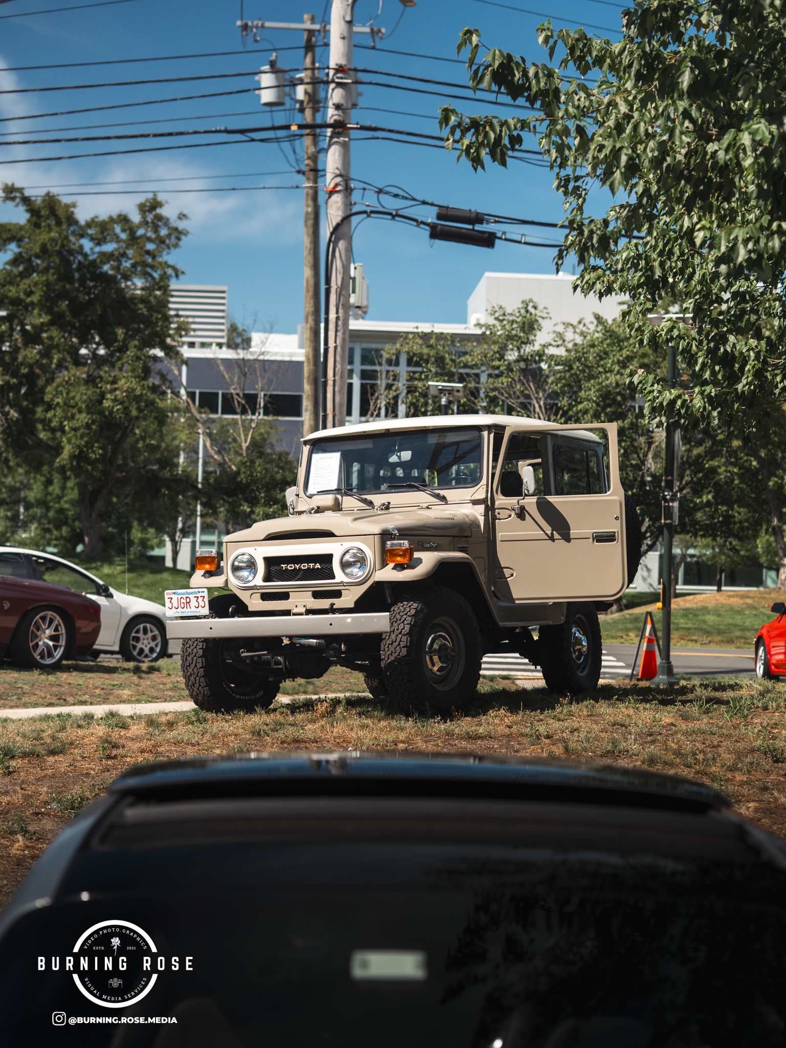 Photo taken from inside a car, showing a beige Toyota Land Cruiser parked on the grass, with other cars and trees in the background under a blue sky.