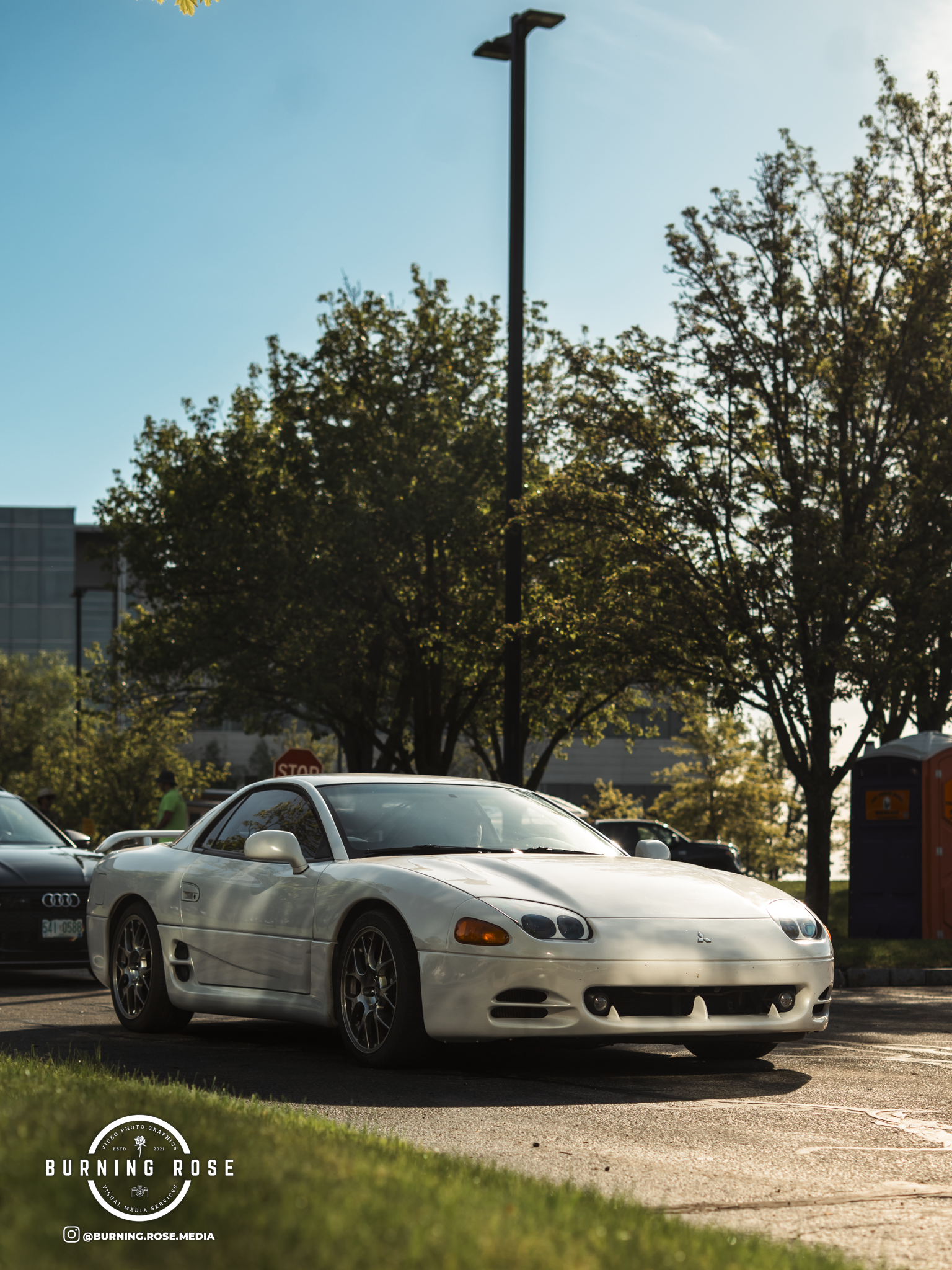 A white sports car parked in a parking lot with trees, other cars, a stop sign, and a purple vending machine in the background on a sunny day.