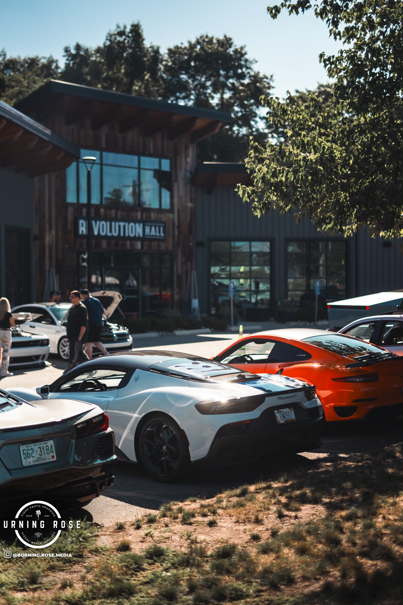 Several luxury sports cars parked in front of a modern wooden building labeled 'Revolution Hall.' People are walking and gathering near the cars, with trees and a clear blue sky in the background.