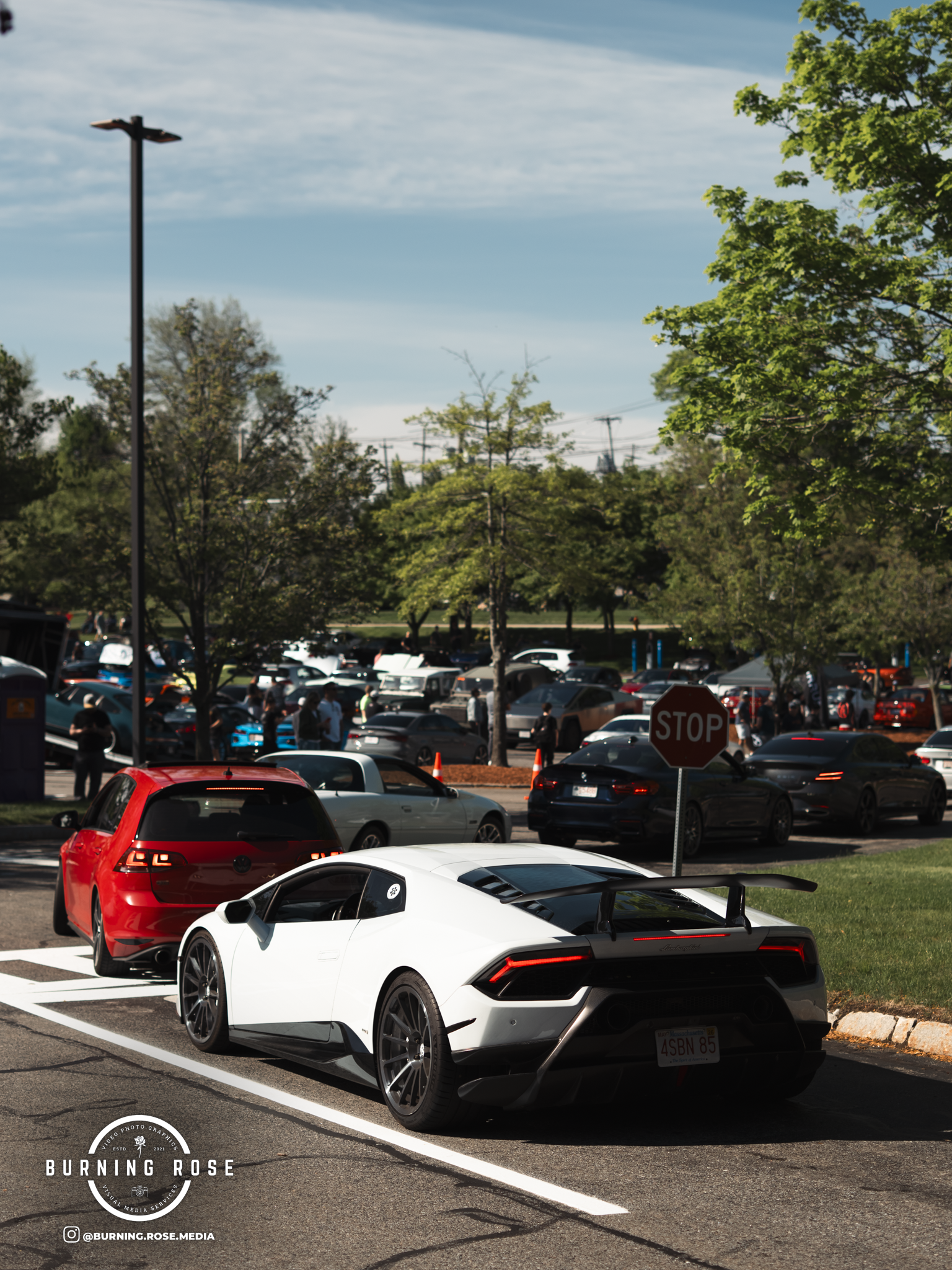 A white Lamborghini sports car parked in a parking lot with other vehicles, trees, a stop sign, and a clear blue sky in the background.