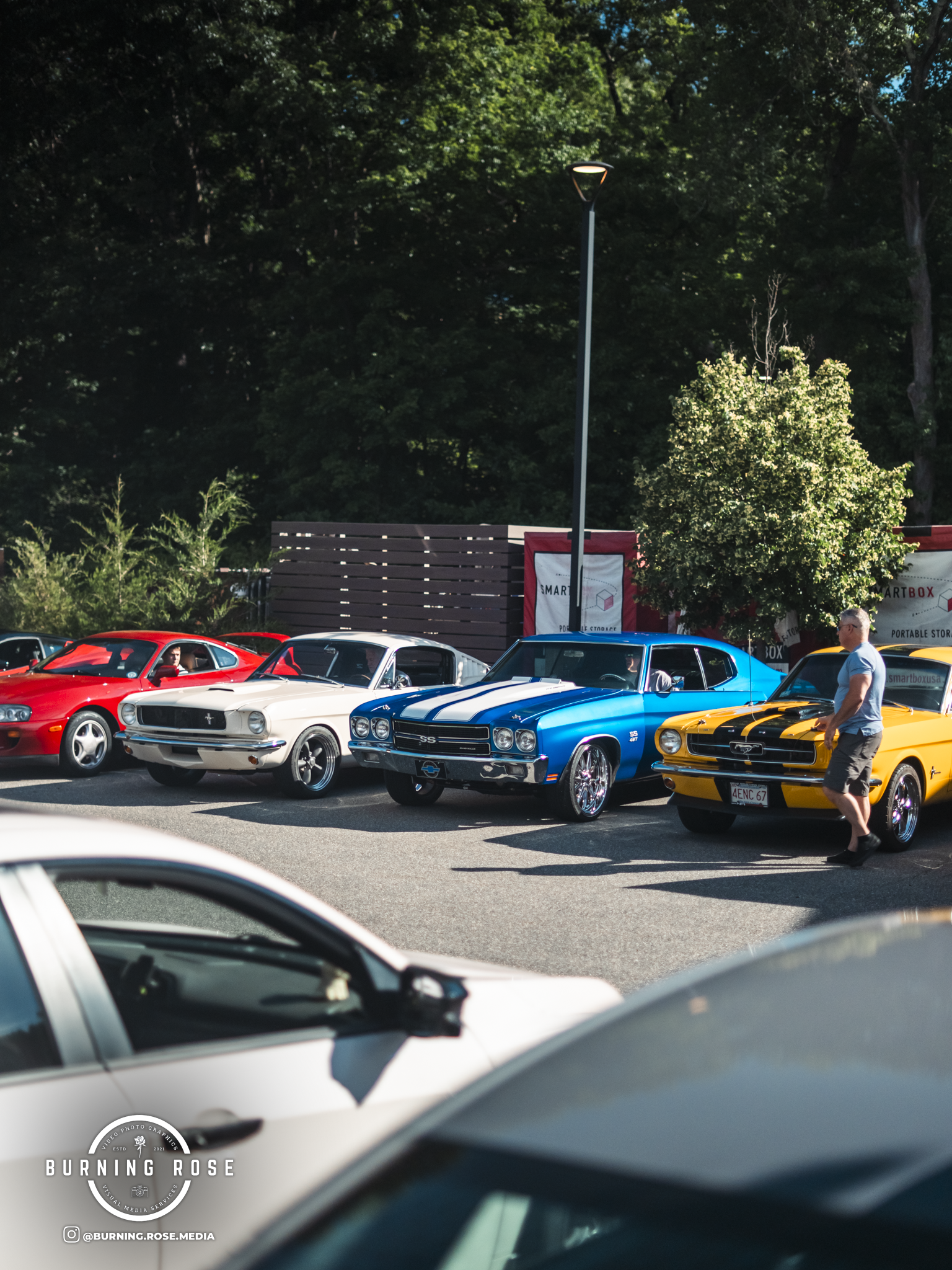 Several classic cars parked in a parking lot with trees and a fence in the background, and a man walking beside a yellow car.