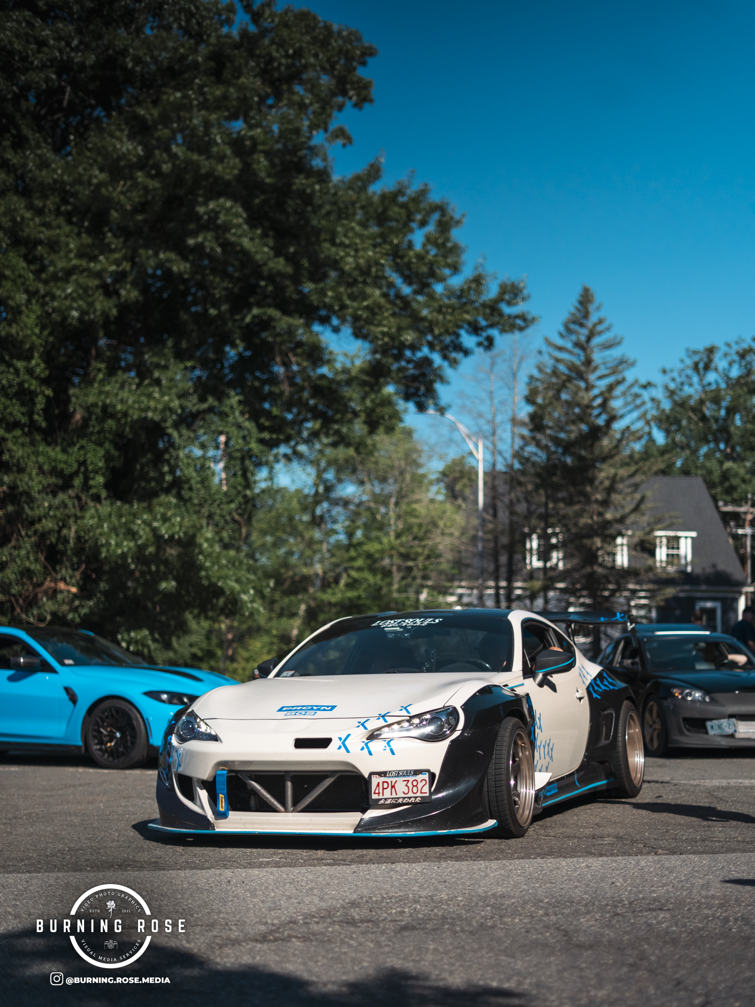 A white and black sports car with blue accents parked among other cars in an outdoor setting with trees and houses in the background.