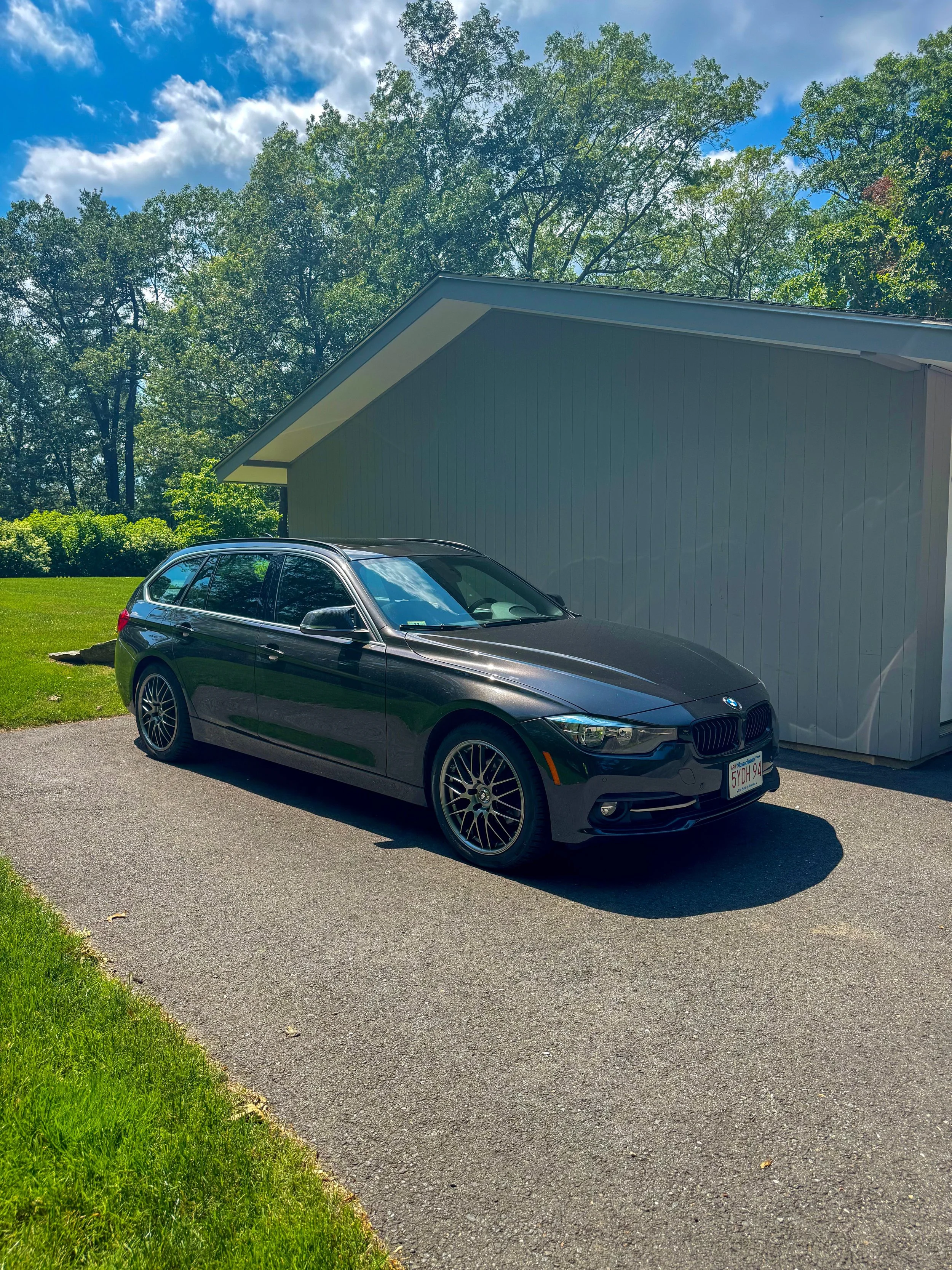 A black BMW station wagon parked on a driveway beside a light gray building, with green grass and trees in the background under a partly cloudy sky.
