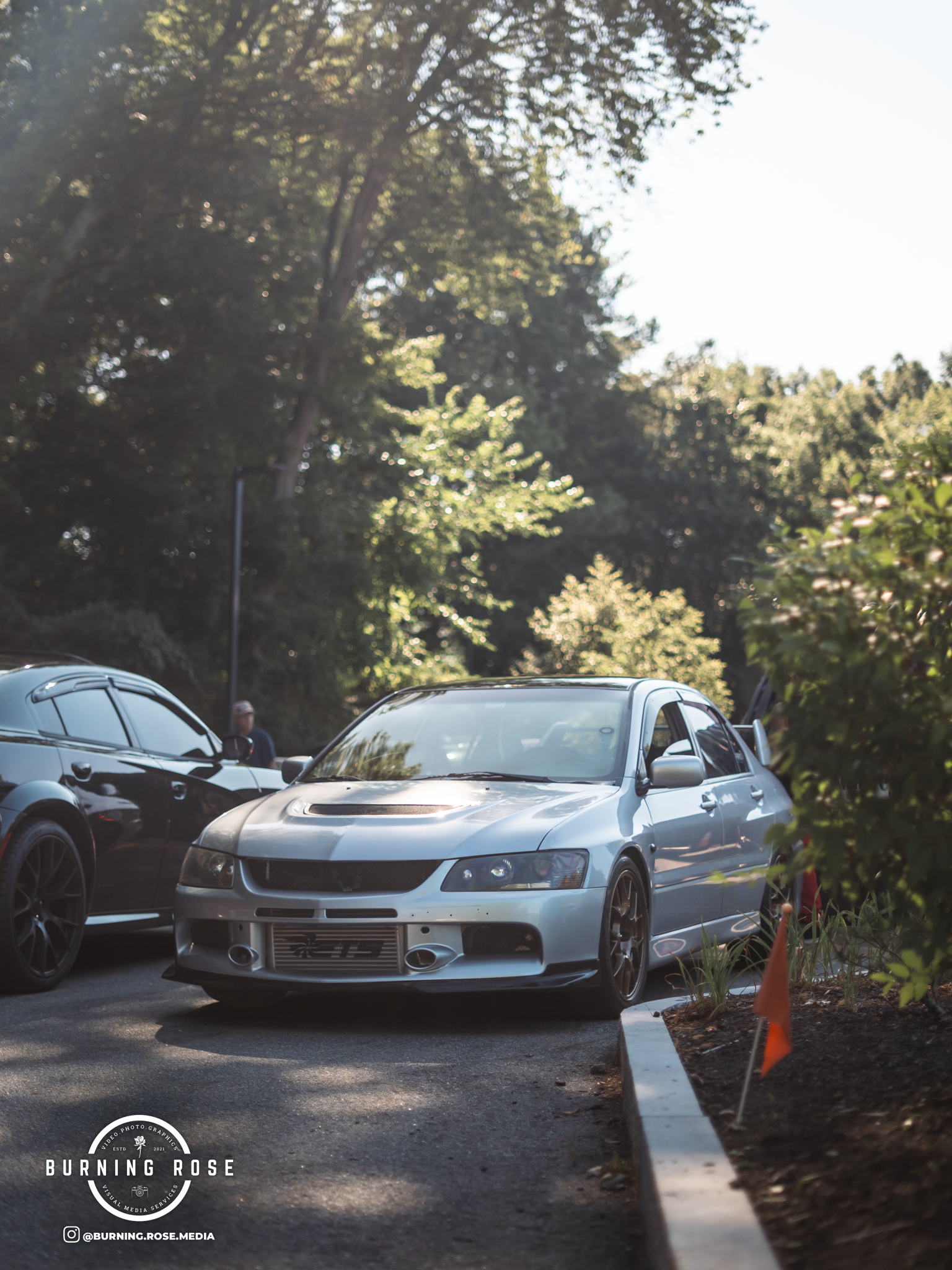 A silver sports car parked in a parking lot surrounded by trees and greenery, with a person standing nearby. The sunlight filters through the leaves, creating a bright background.