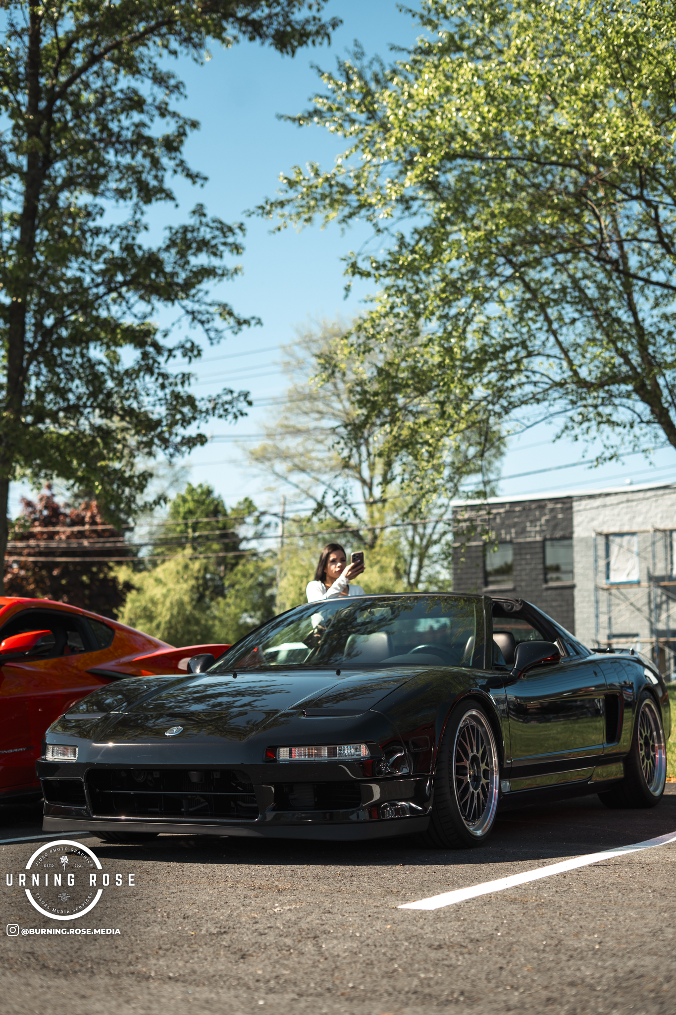 A woman with long dark hair in a white shirt standing next to a black sports car, taking a photo with her phone at a car show. There are red and black cars parked in the background, trees with green leaves, a brick building, and blue sky.
