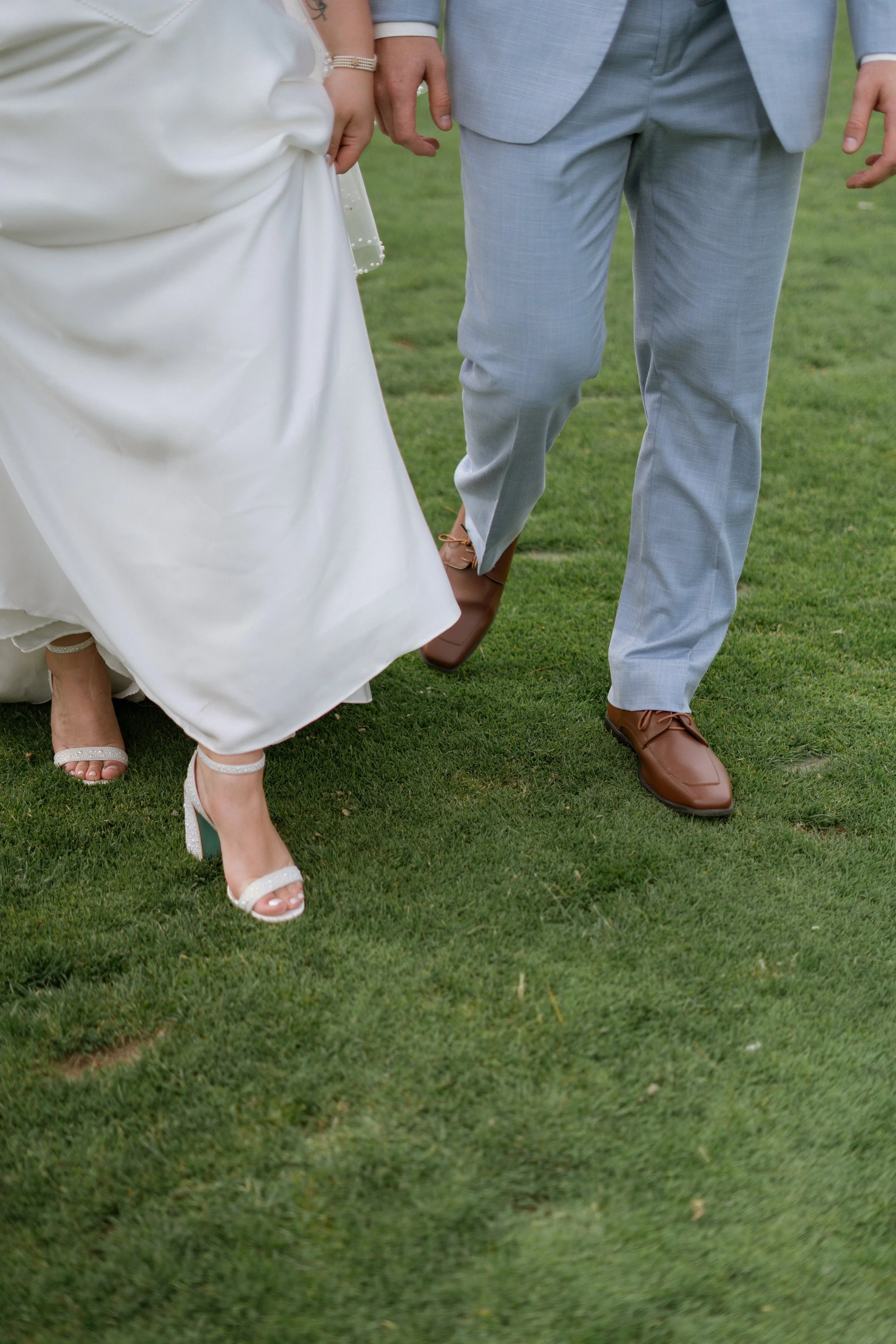 Close-up of a bride and groom holding hands walking on grass. The bride is wearing a white wedding dress and high-heeled sandals, and the groom is wearing a light gray suit with brown shoes.