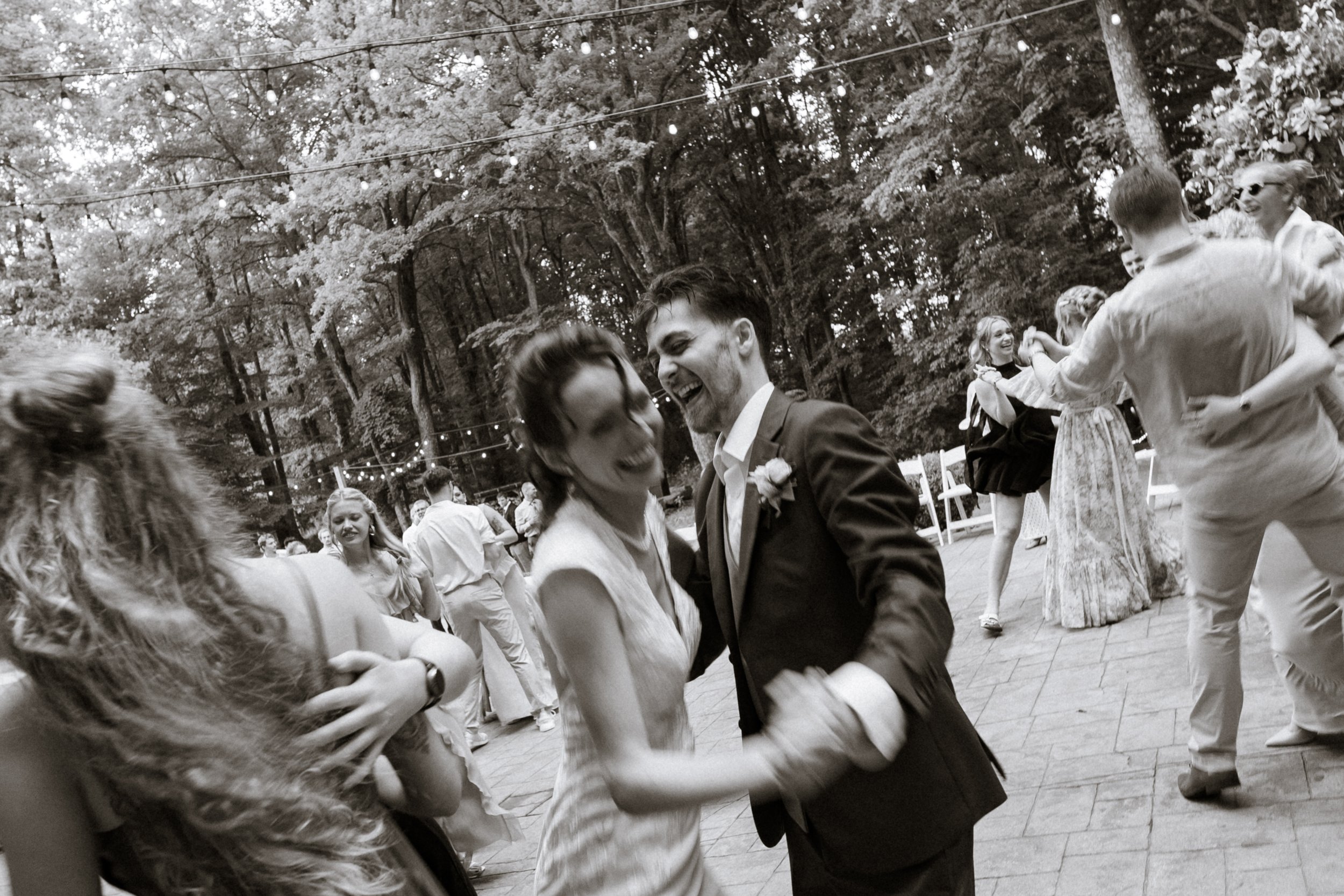People dancing and celebrating outdoors at a party with string lights overhead in a wooded area.